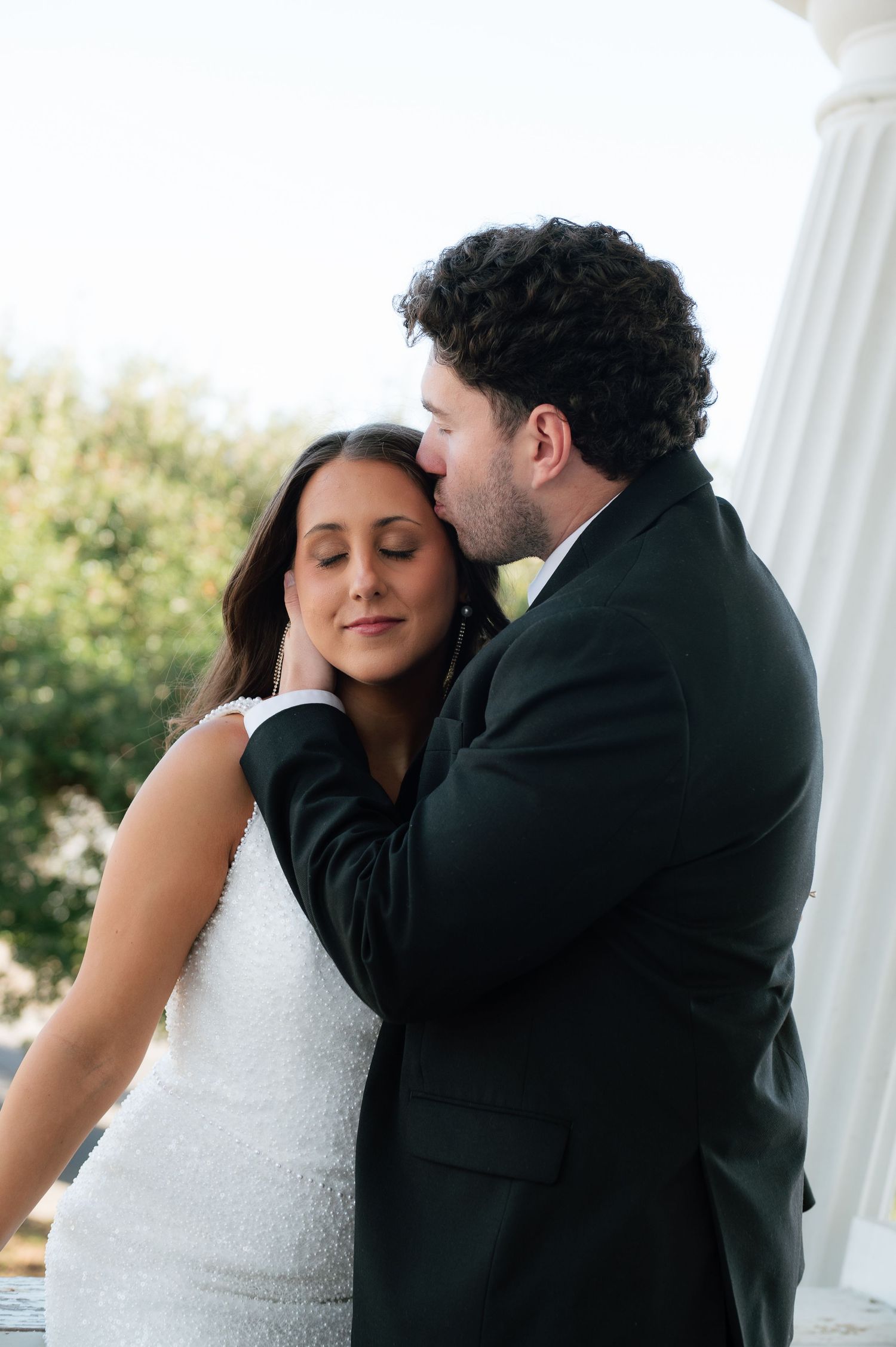 A groom in a black suit tenderly kisses his bride on the cheek as she smiles with closed eyes in a white dress.