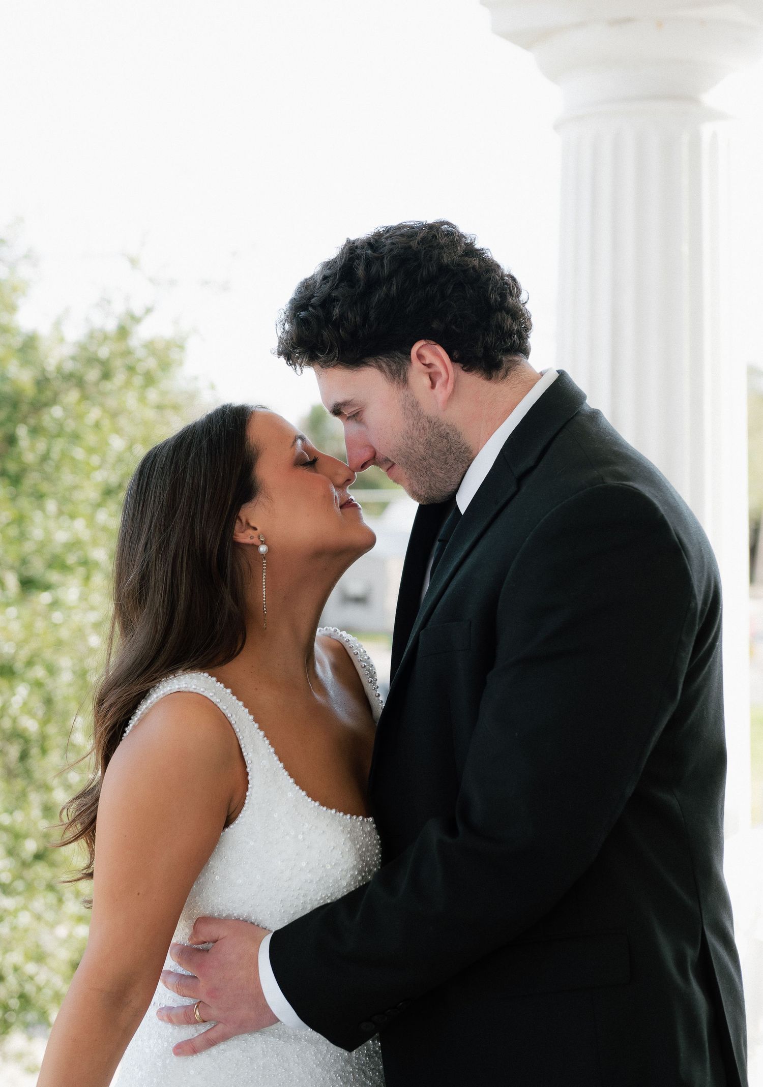 A bride and groom share a romantic almost-kiss moment near white columns, dressed in wedding attire.