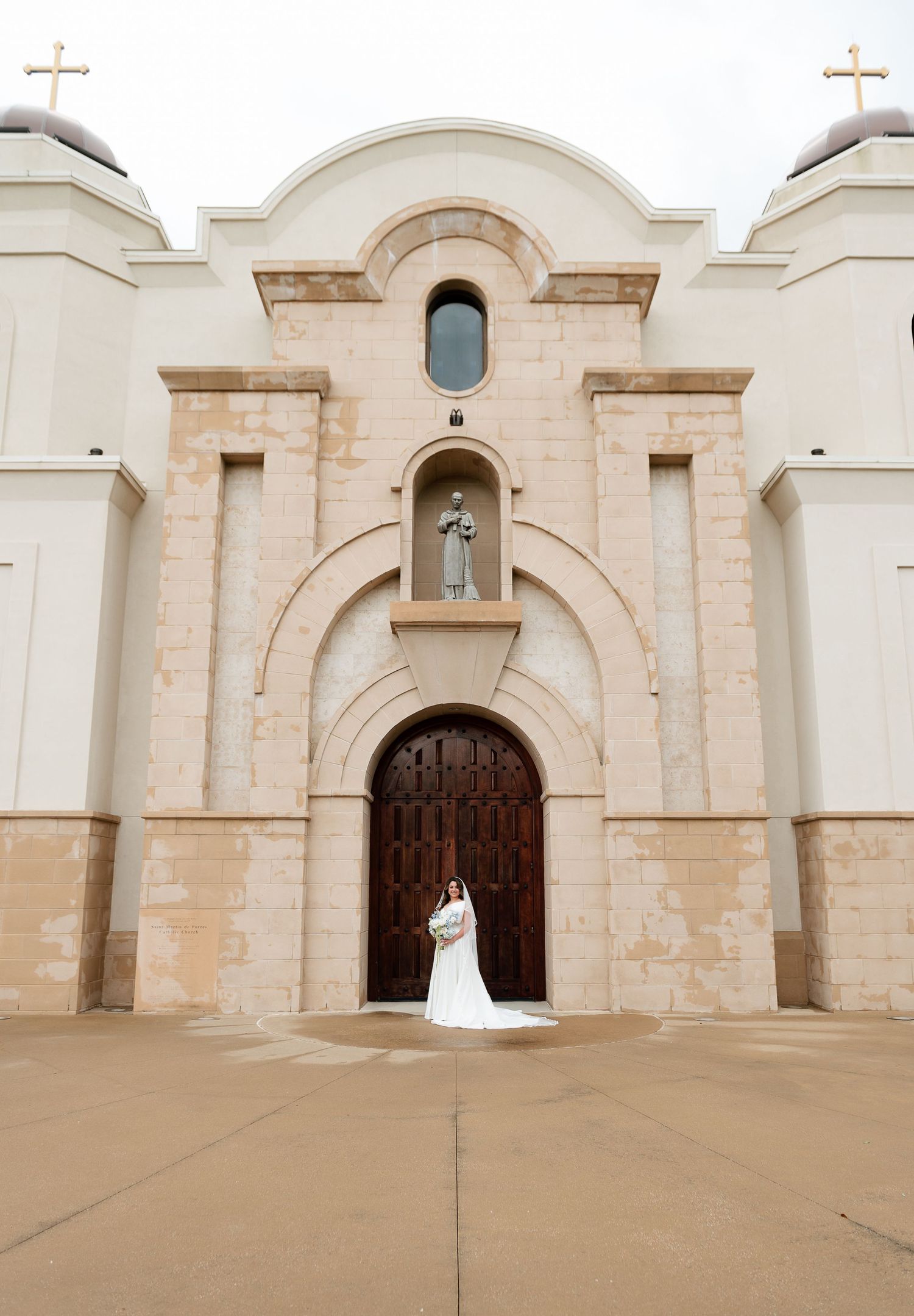 Bride in white gown standing before ornate church entrance with arched wooden doors and religious statue.