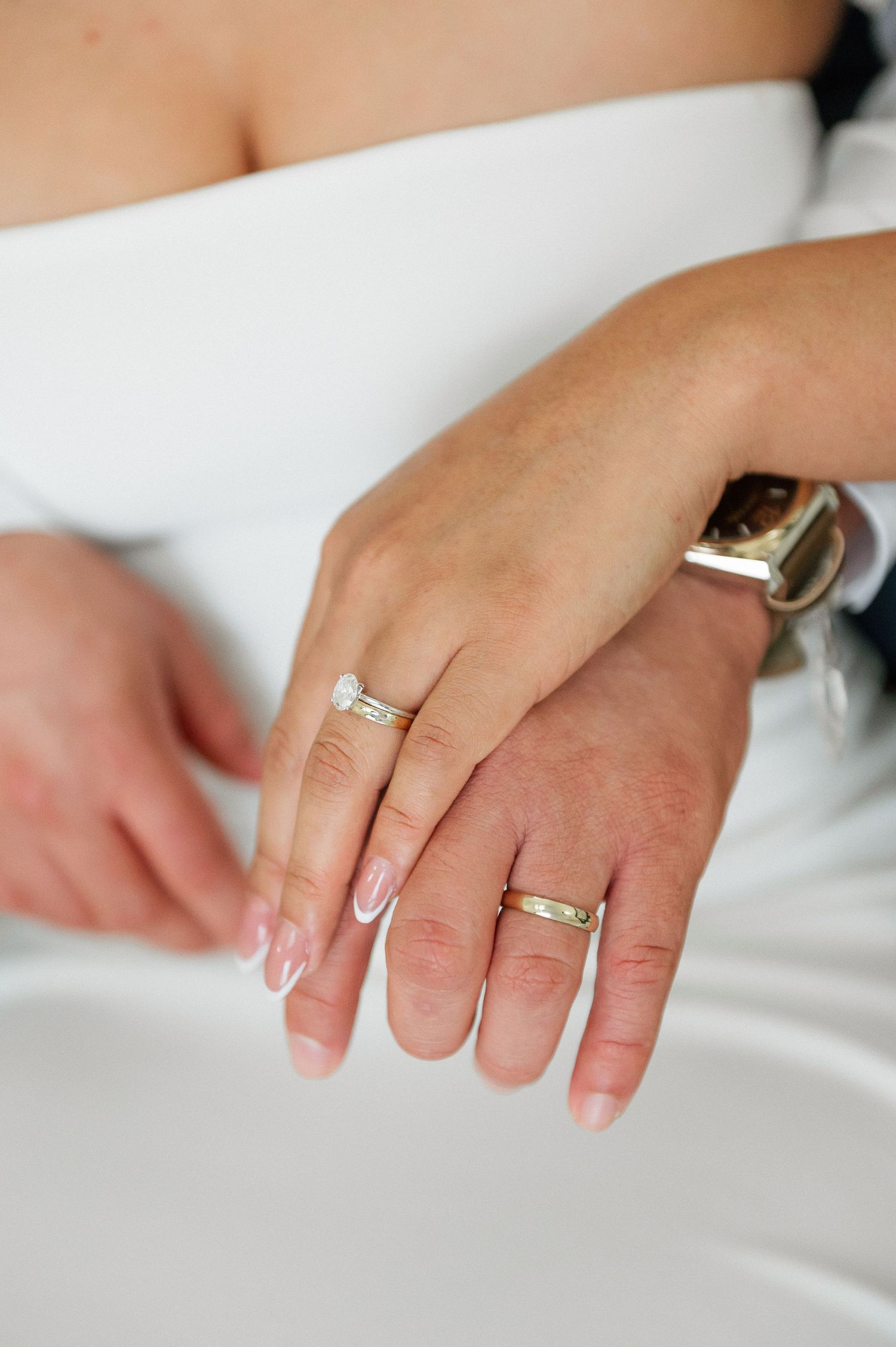 Newlywed couple displaying wedding rings on ring fingers, bride in white dress with groom wearing a watch.
