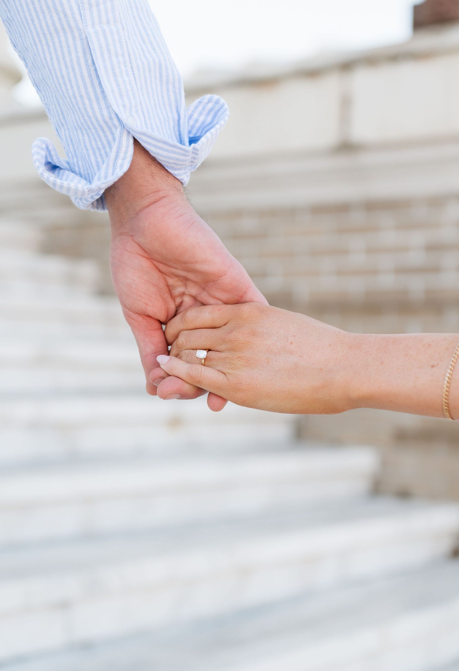Couple holding hands on marble steps, woman wearing an engagement ring, man in light blue shirt.