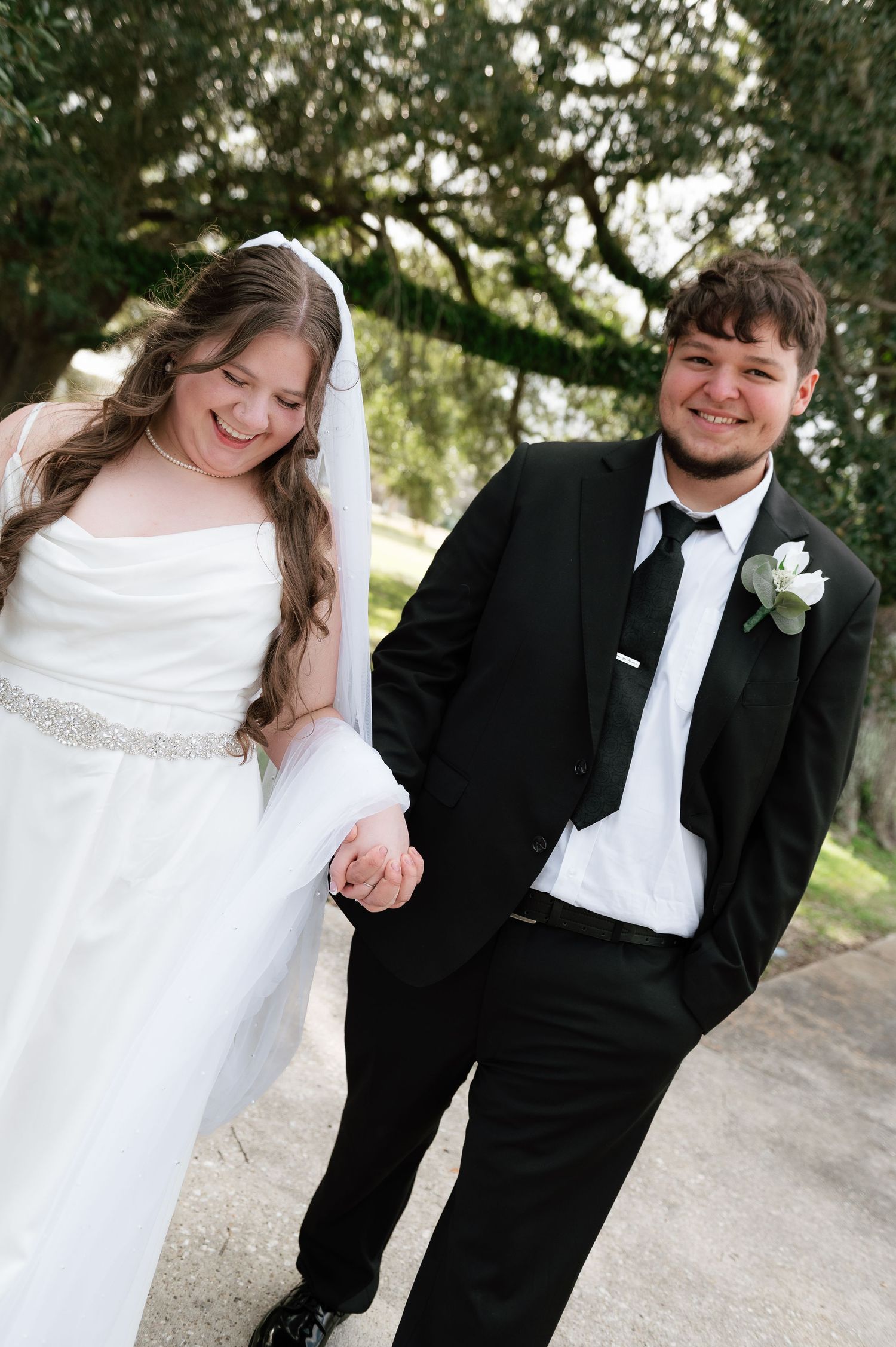 Smiling bride in white gown and veil holds hands with groom in black tuxedo outdoors under oak trees.