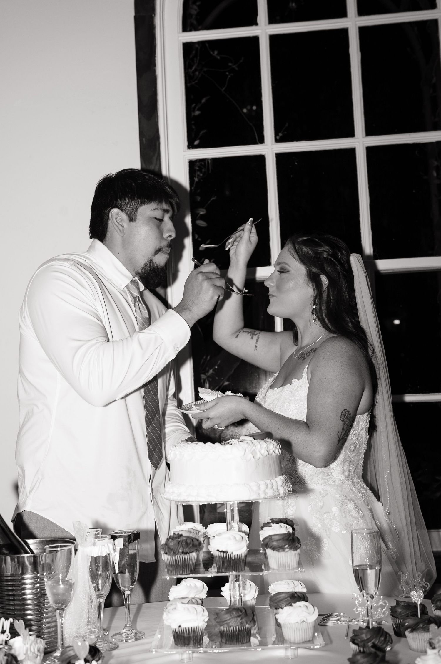 Black and white photo of bride and groom feeding each other wedding cake at reception with cupcake display.