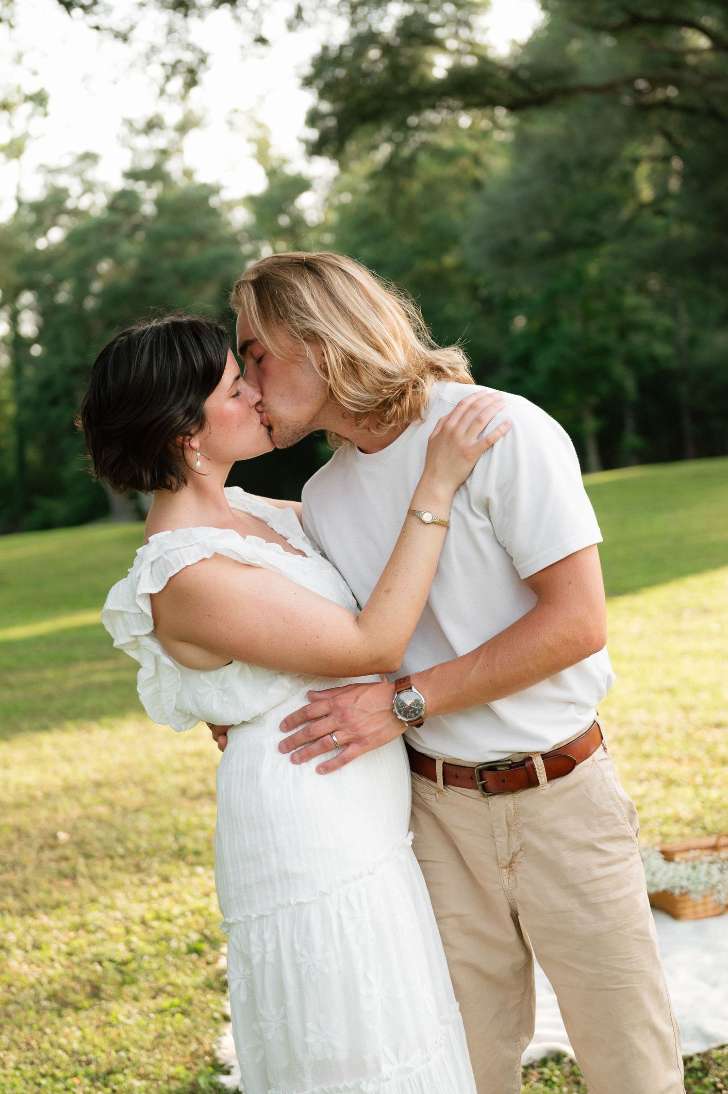 A couple shares a romantic kiss outdoors on a sunny day, dressed in white and khaki attire on a lush green lawn.