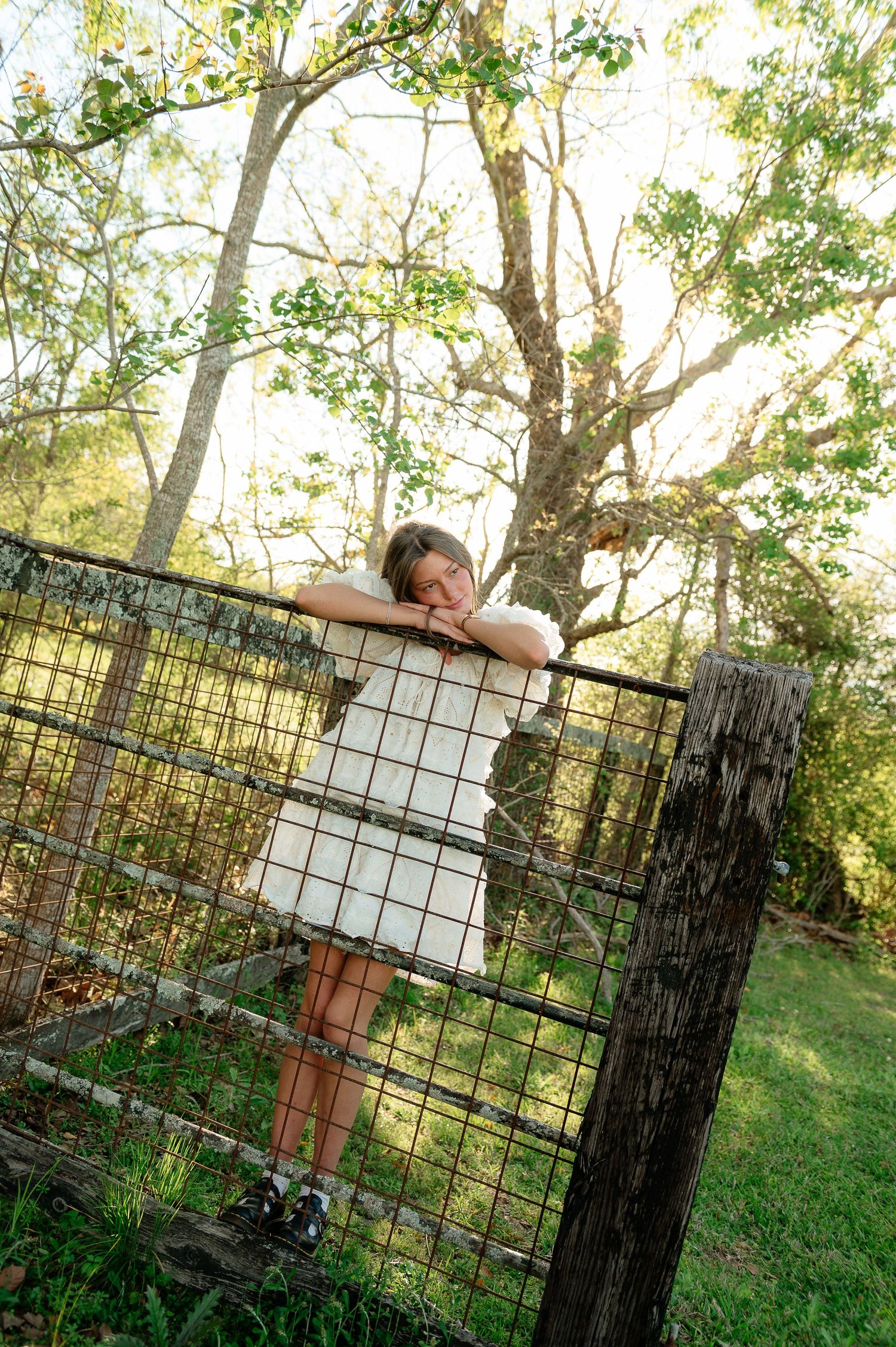 Young woman in white dress leaning on a rustic wire fence surrounded by sunlit trees outdoors.