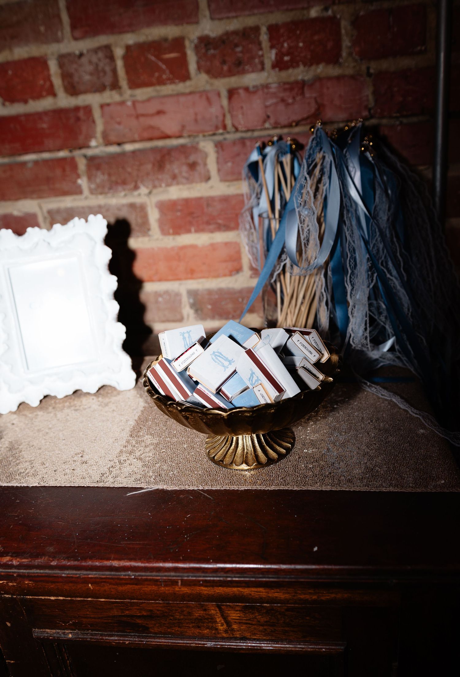 Gold pedestal bowl filled with wedding favor matchbooks and ribbon wands on rustic wooden dresser.