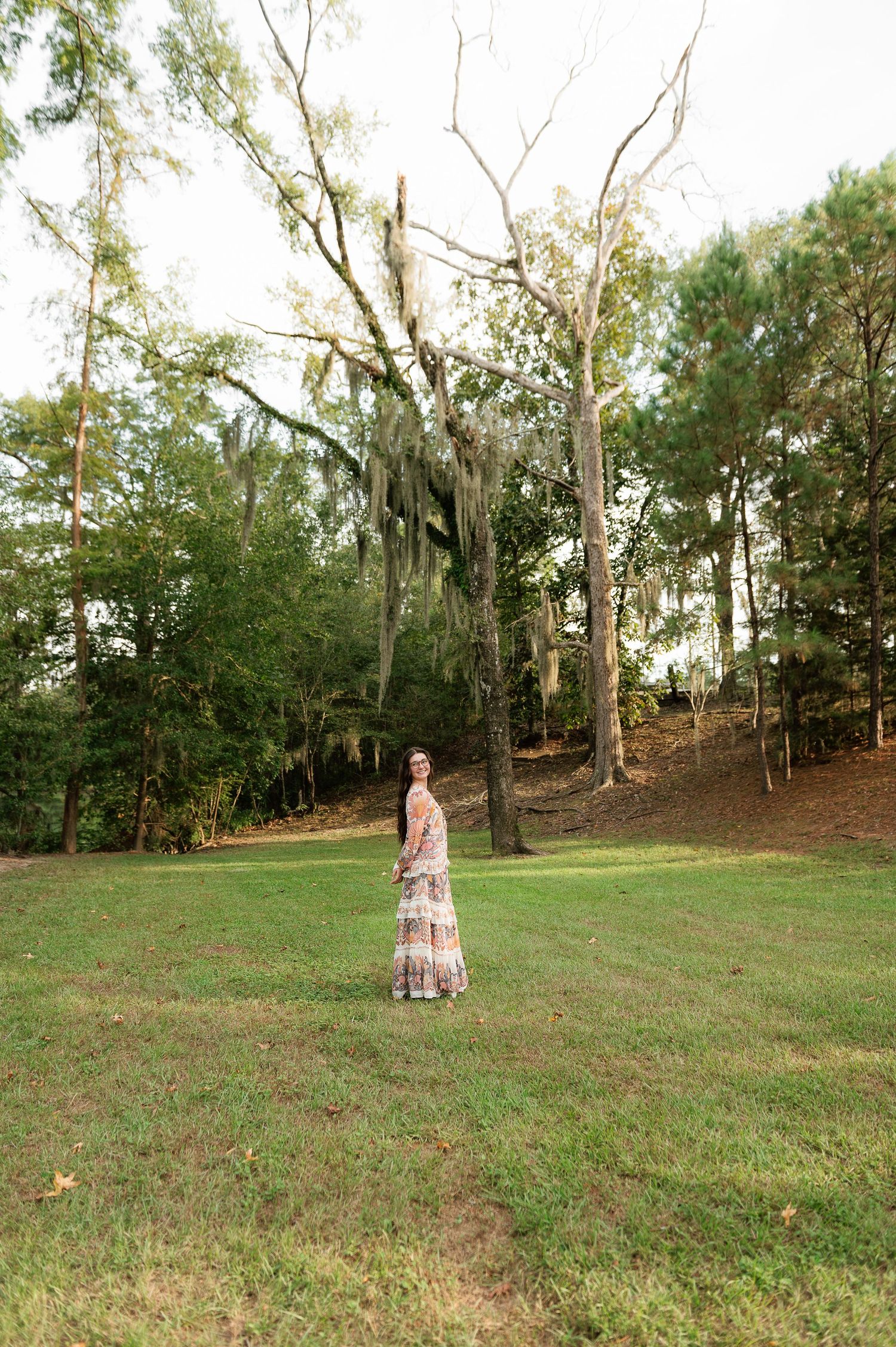 Woman in a long floral maxi dress stands on a grassy lawn surrounded by tall trees draped in Spanish moss.