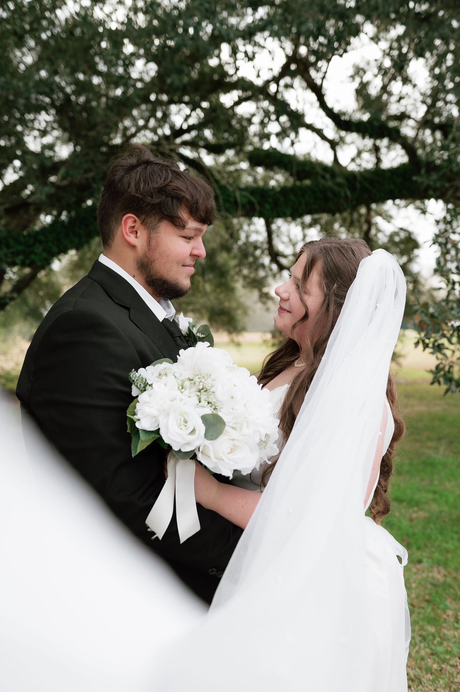A bride and groom share a tender moment outdoors, she holds white roses bouquet, wearing veil, he wears black suit.