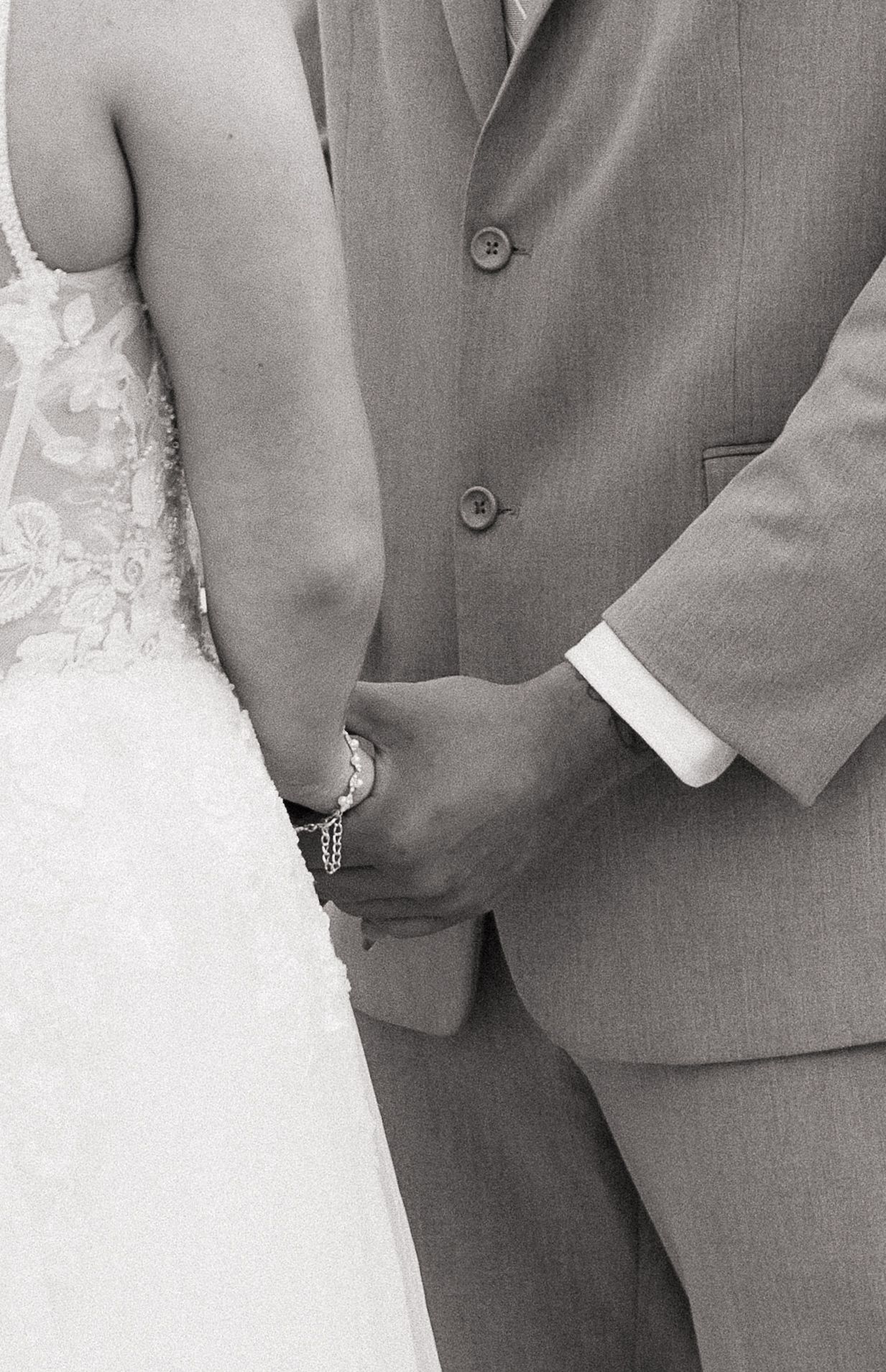 Black and white wedding photo of bride in lace gown and groom in gray suit holding hands.