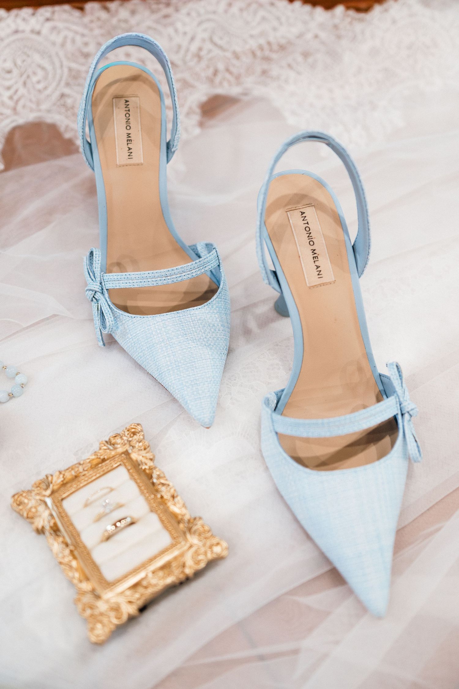 Light blue slingback heels with bows beside a gold frame with wedding rings on white tulle.