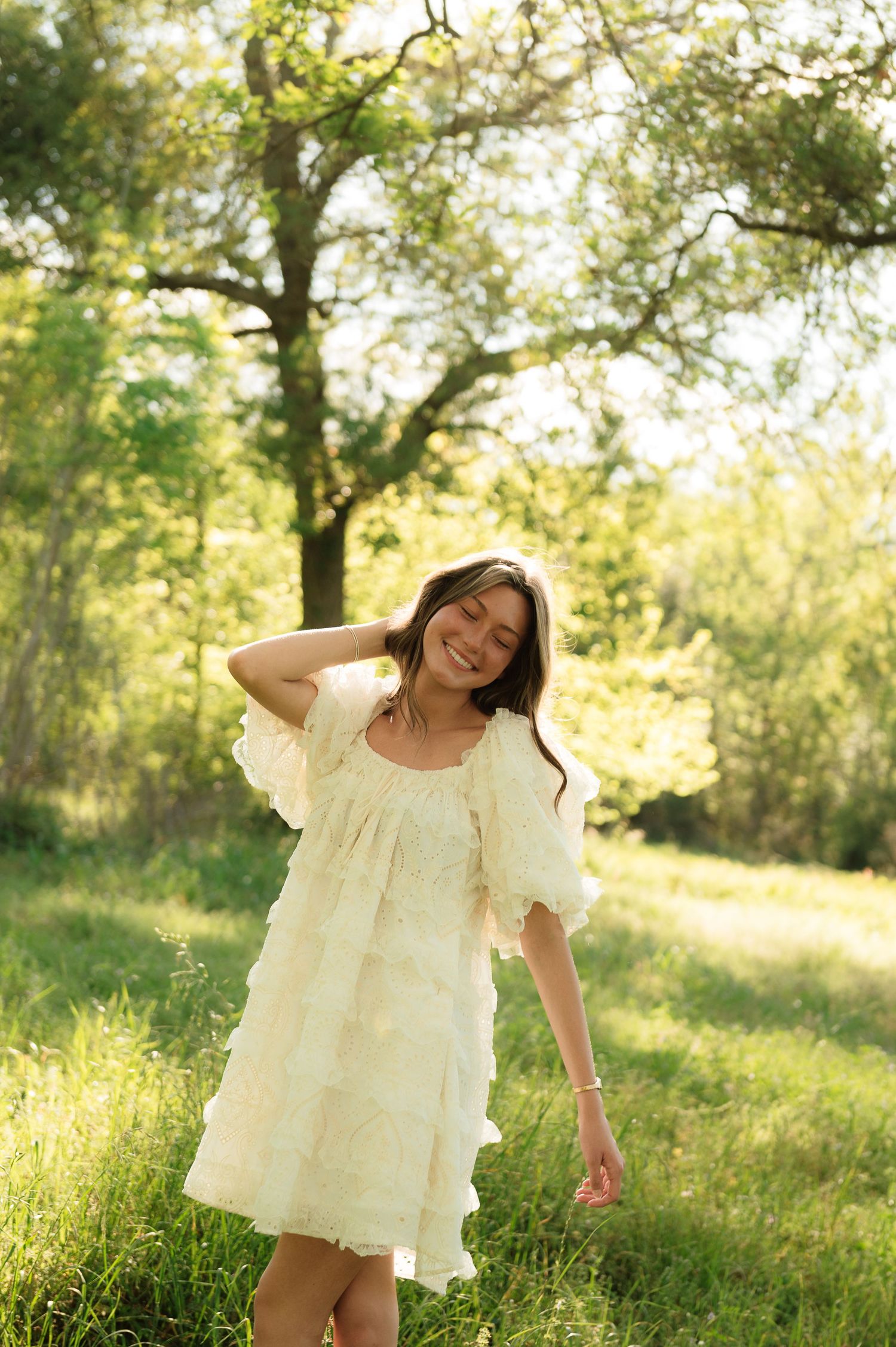 Young woman in white ruffled dress smiling outdoors in sunny green meadow with trees.