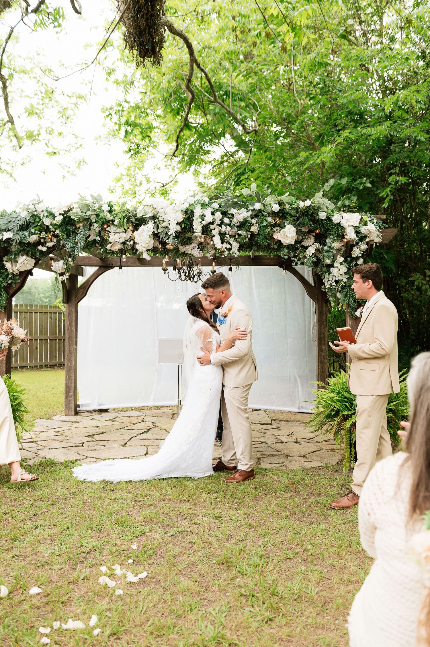 Bride and groom share first kiss under floral arch at outdoor garden wedding ceremony with white blooms.