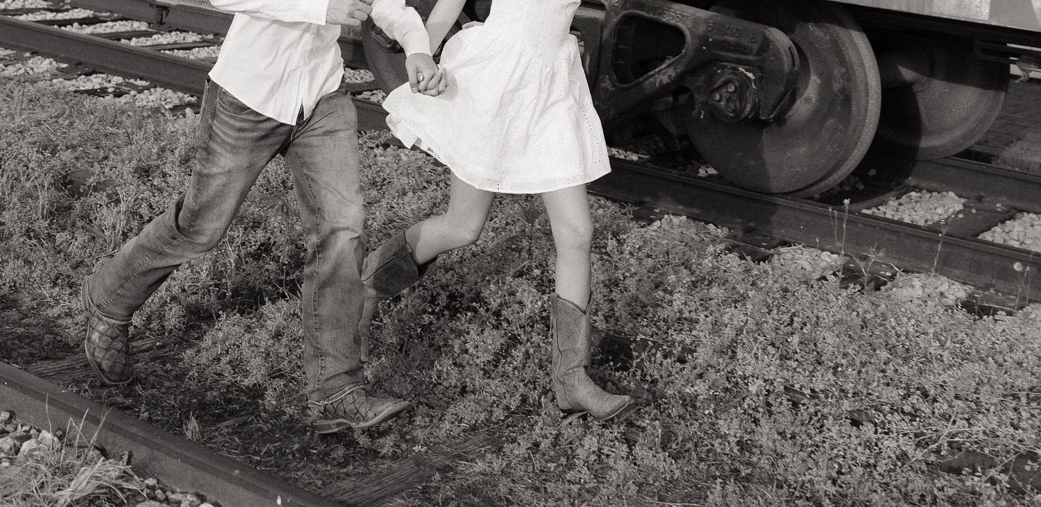 Couple in cowboy boots holding hands near railroad tracks in black and white photo.