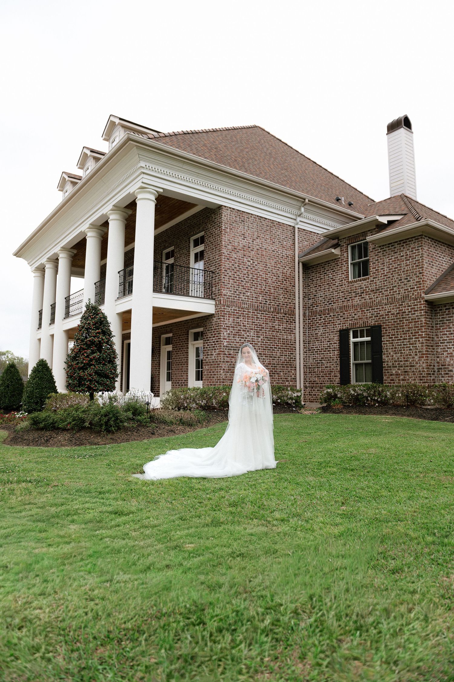 Bride in white wedding gown with long veil stands on green lawn before elegant brick mansion with white columns.