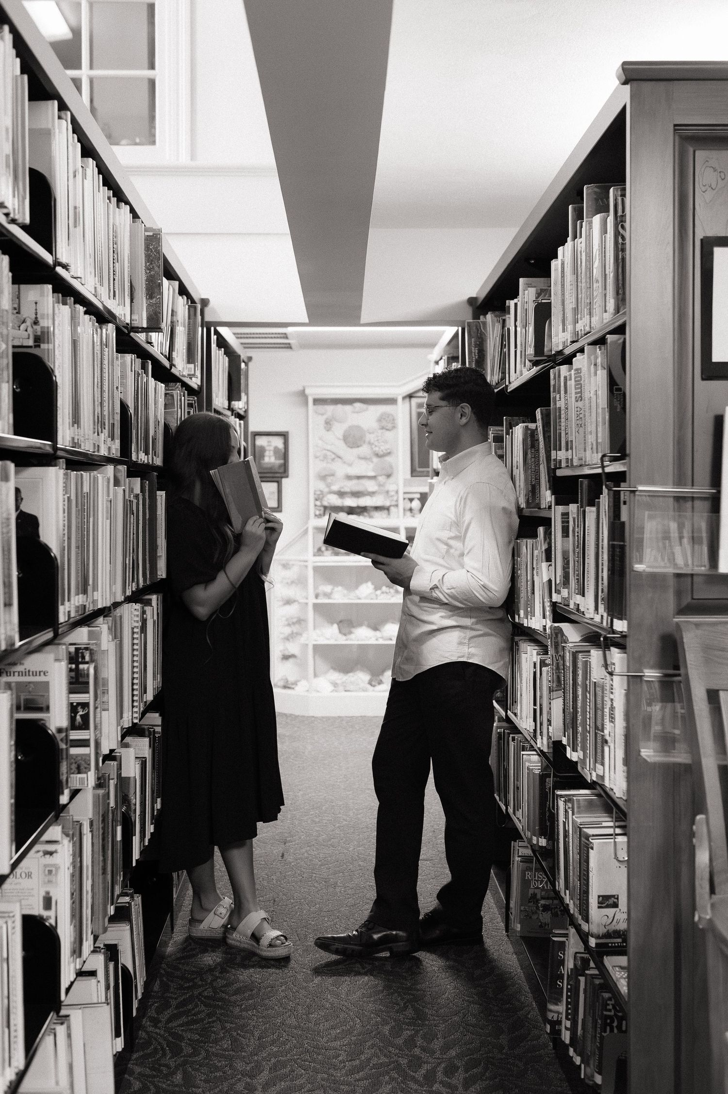 Two people stand between library bookshelves, reading books in a black and white photo.