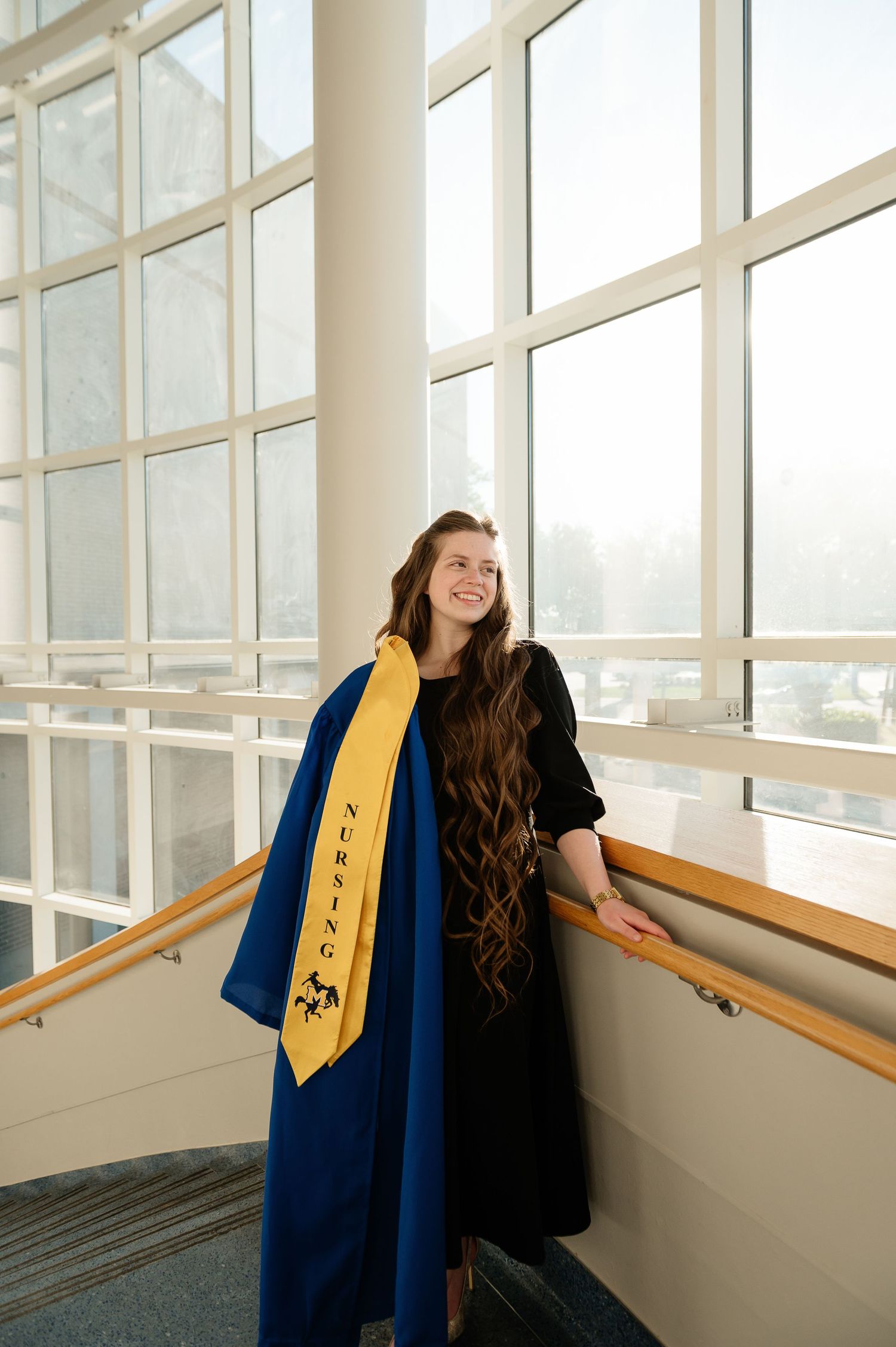 Smiling nursing graduate in blue gown and gold stole poses by large windows in a bright modern building.