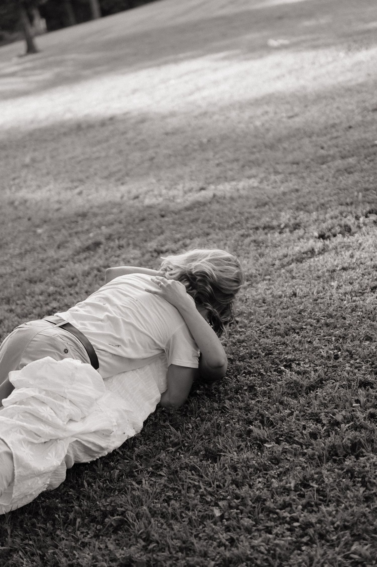 Black and white photo of a woman in a white dress lying face down on a grassy lawn outdoors.