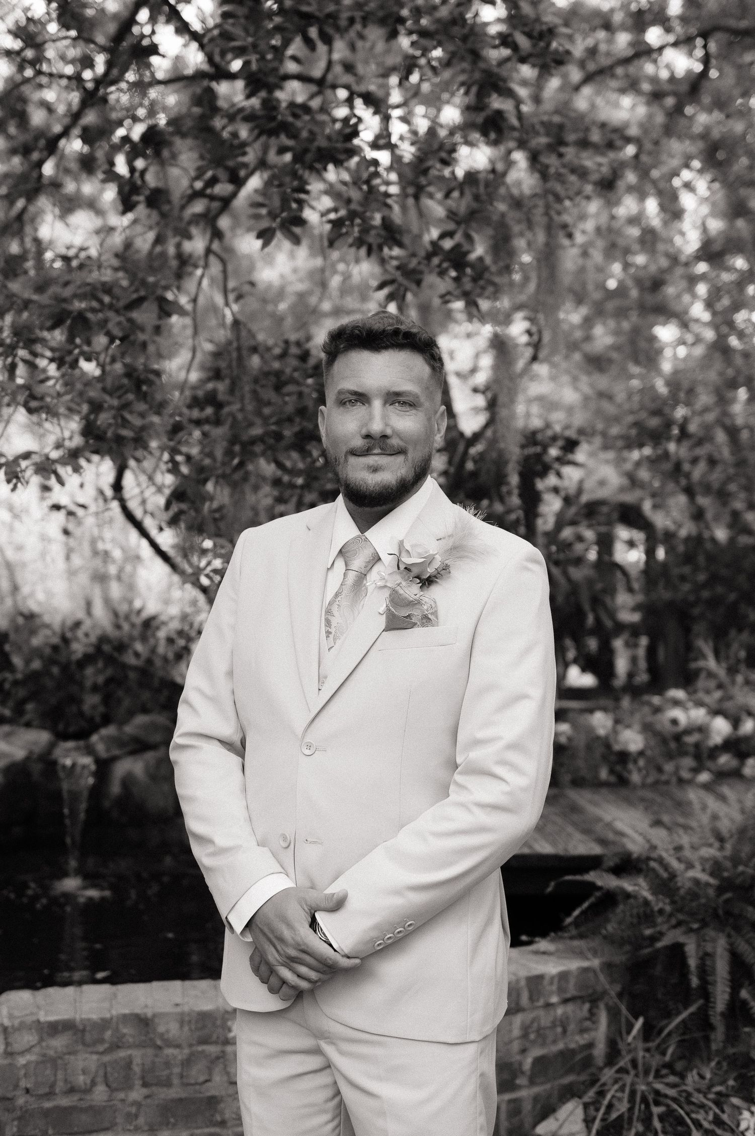 Groom in white suit with floral boutonniere poses outdoors in lush garden setting, black and white photo.