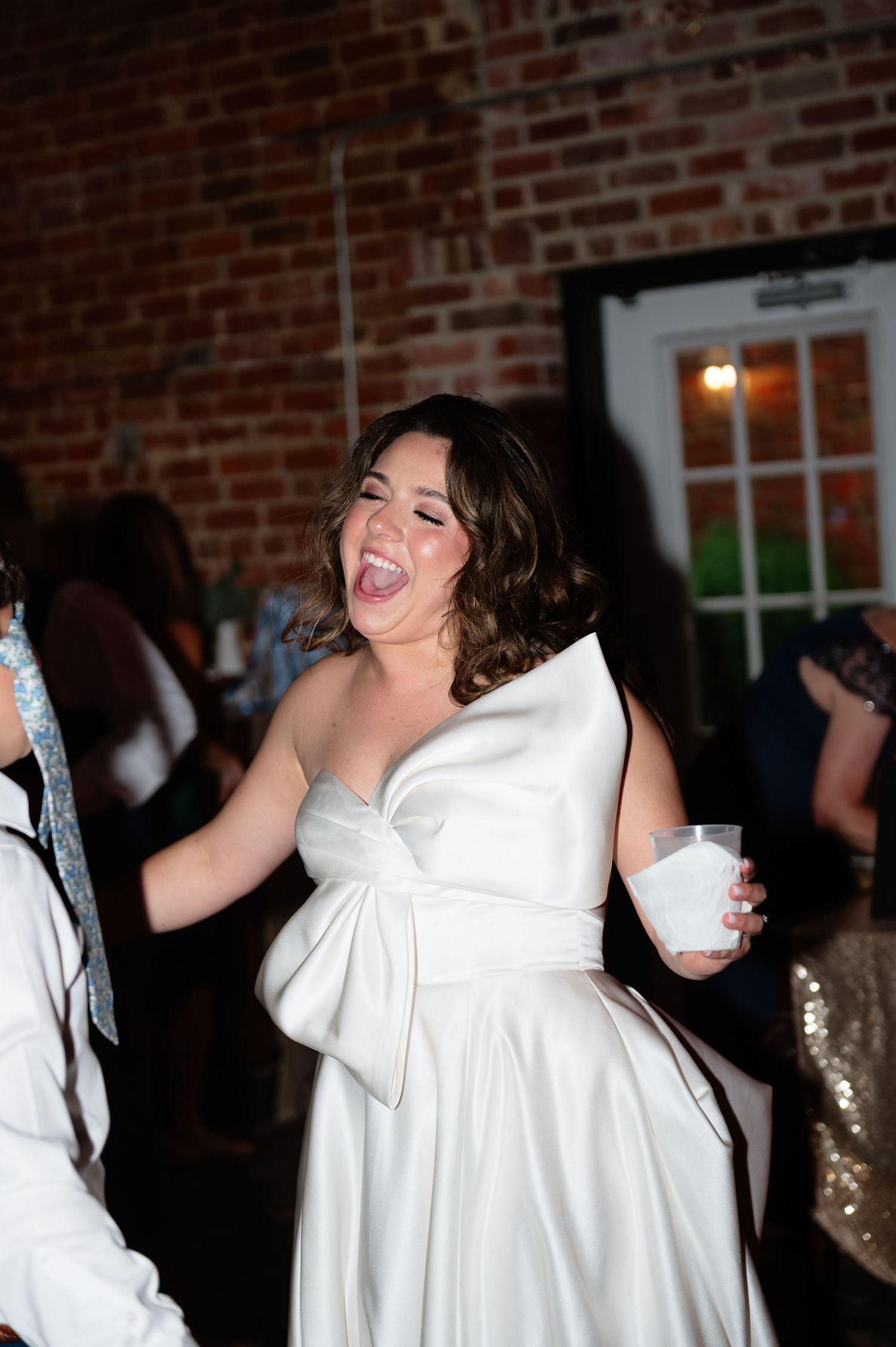 Bride in white satin gown laughing joyfully while dancing at wedding reception.