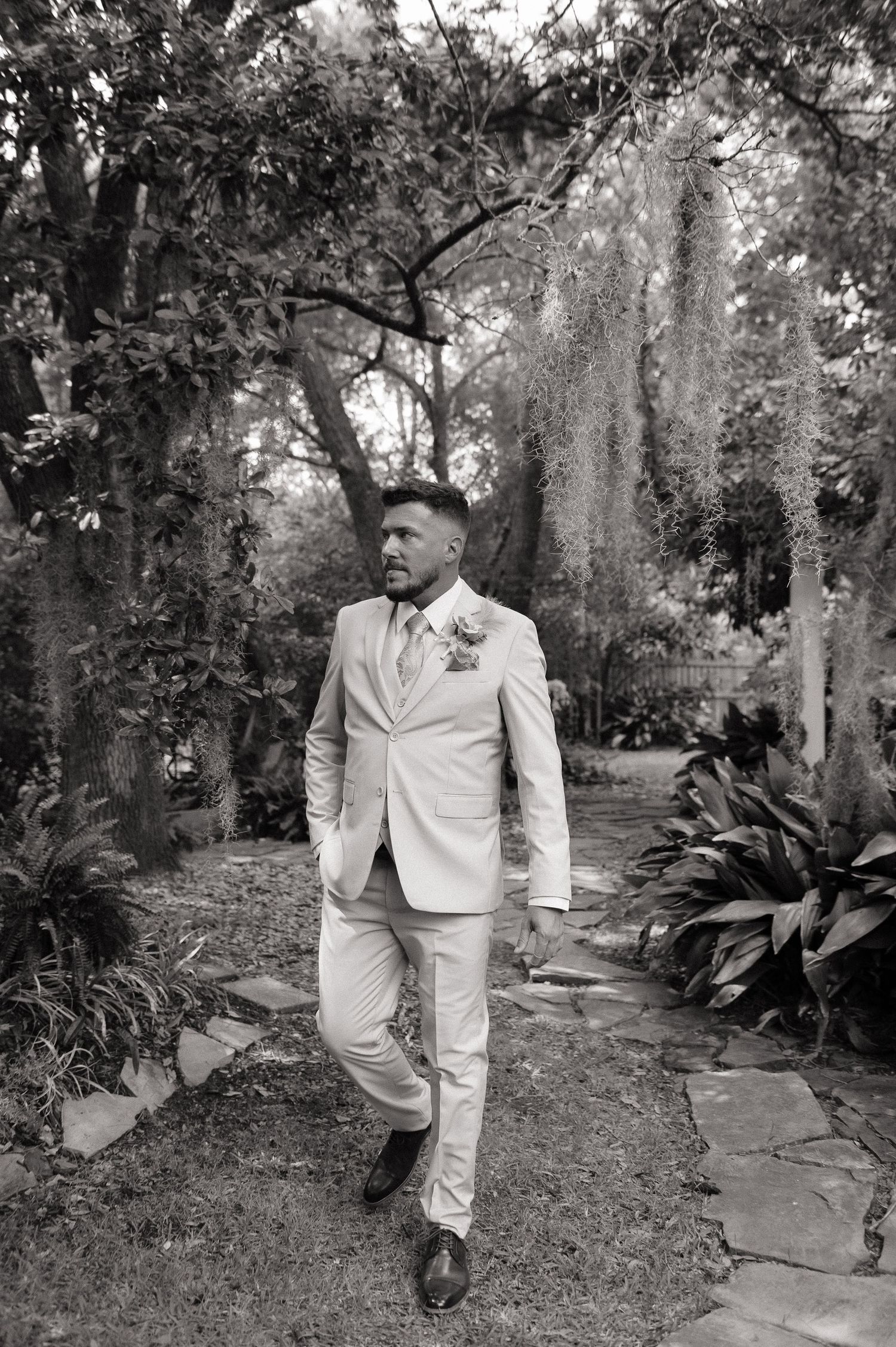 A groom in a light suit with boutonniere walks a garden stone path surrounded by lush trees and Spanish moss.