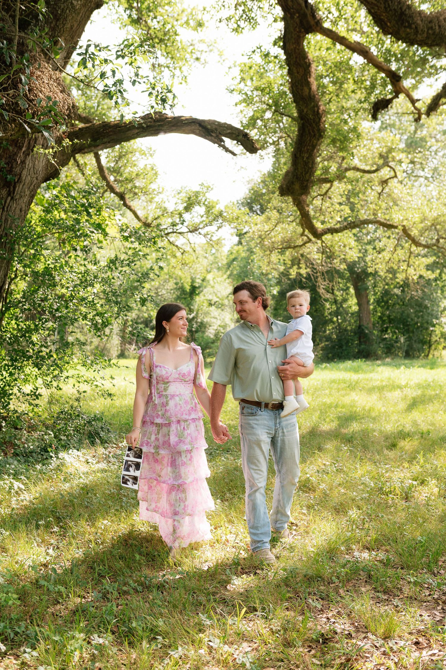 Pregnant woman in pink floral dress walks with man holding toddler through sunny green park.