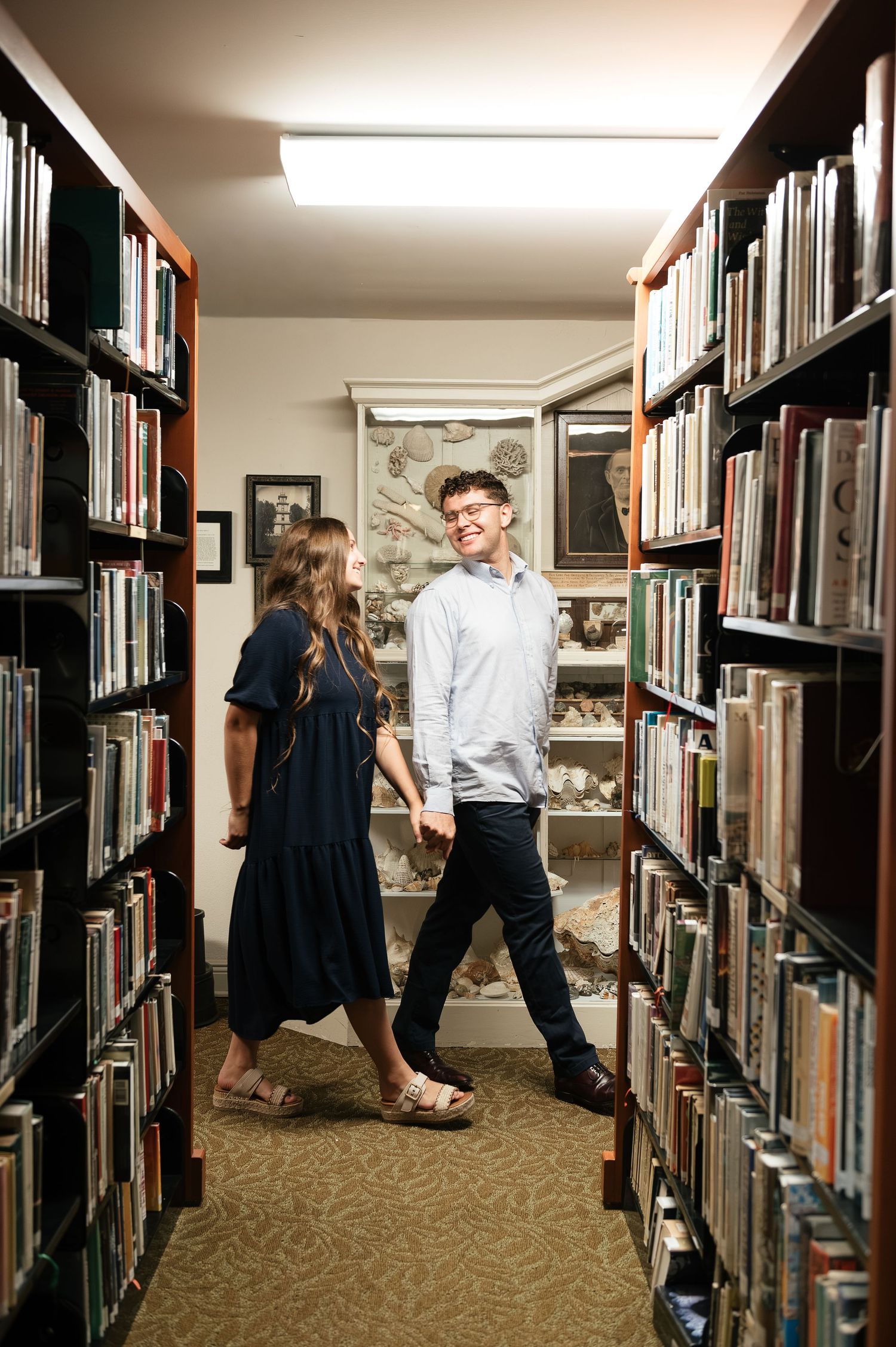 A couple walks hand-in-hand between tall library bookshelves, smiling at each other in a cozy, warmly lit library.
