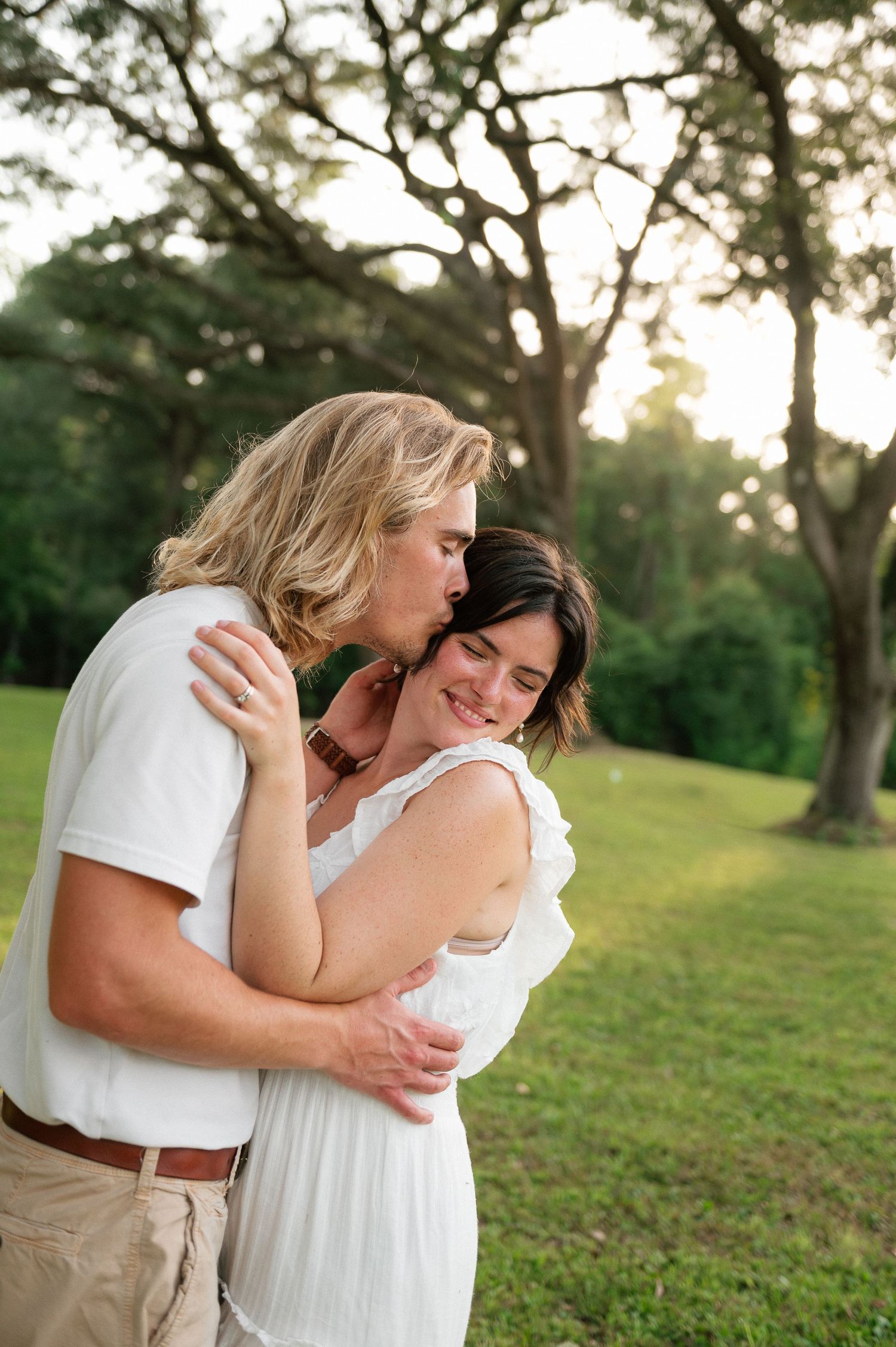 A couple embraces outdoors, man kissing woman's temple while she smiles, surrounded by lush green trees.