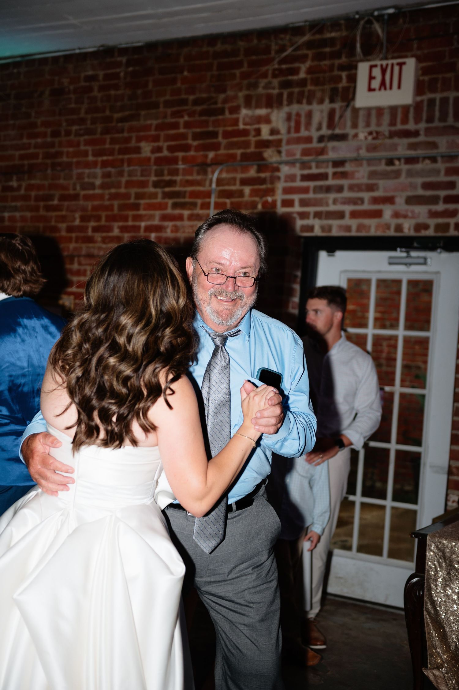 Bride shares father-daughter dance at rustic brick-walled wedding reception venue.