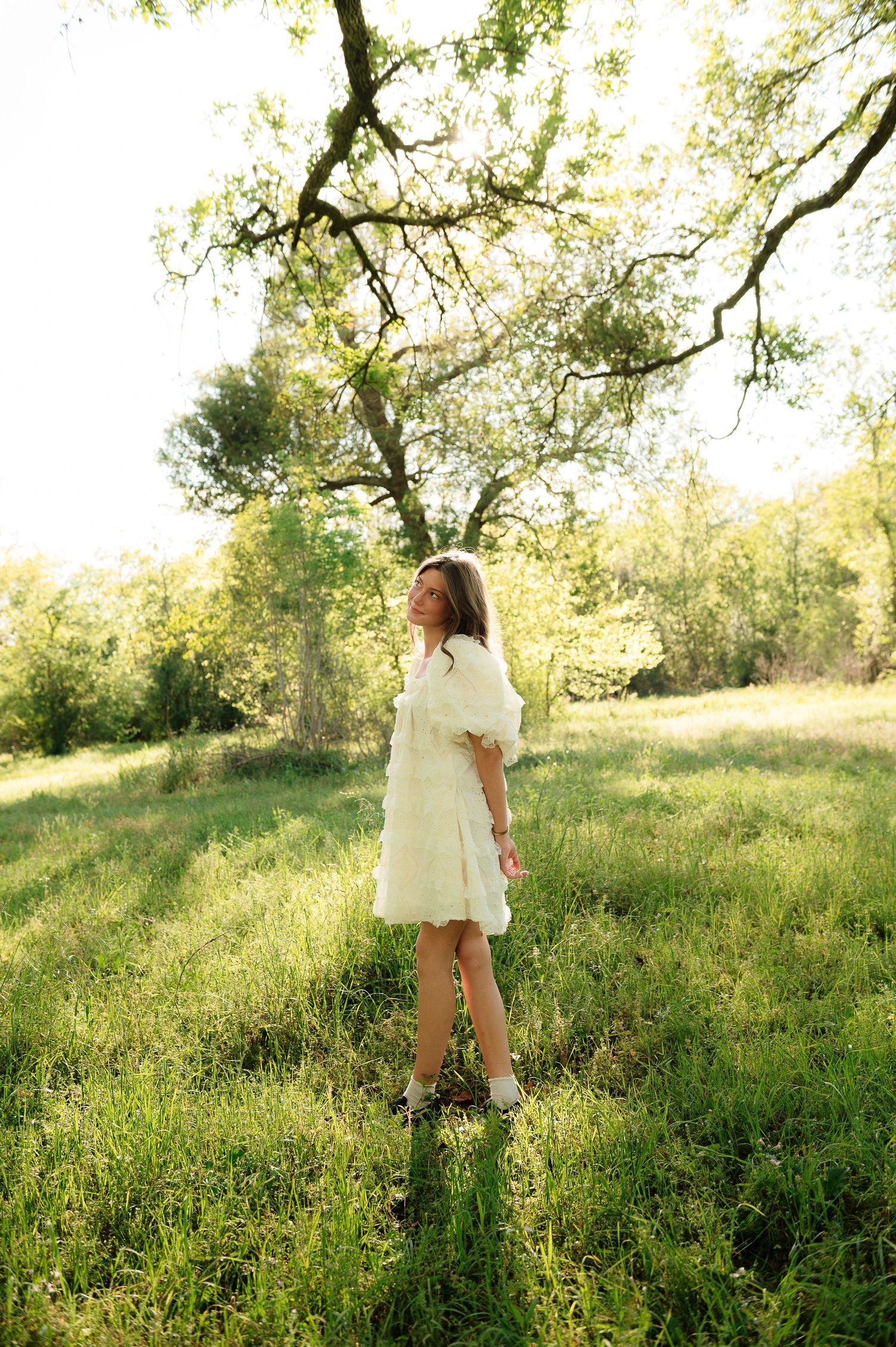 Young woman in white dress standing in sunlit green meadow beneath a large oak tree on a sunny day.