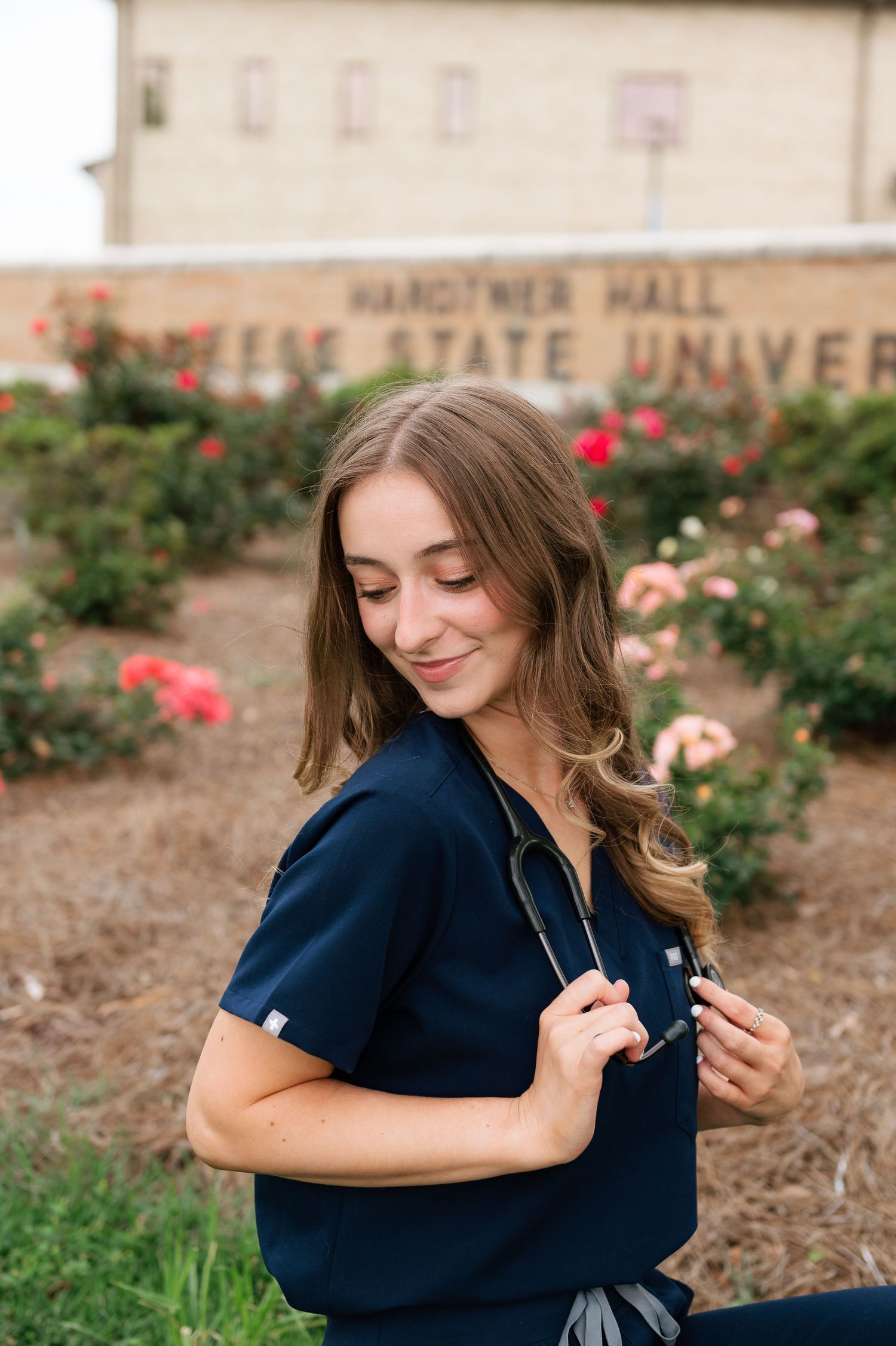 Nursing student in navy scrubs with stethoscope posing in front of a state university building surrounded by roses.