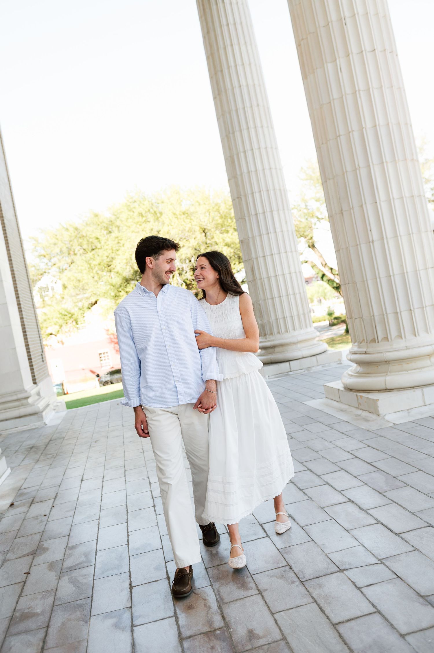 A smiling couple walks hand-in-hand between tall white columns, dressed in light outfits on a sunny day.