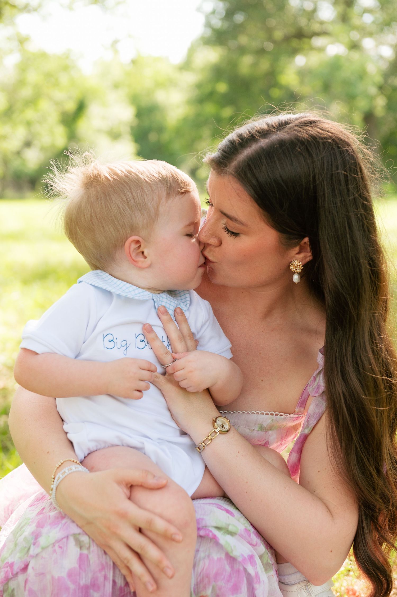 A young mother with long dark hair kisses her blonde toddler boy outdoors in a sunny green park setting.