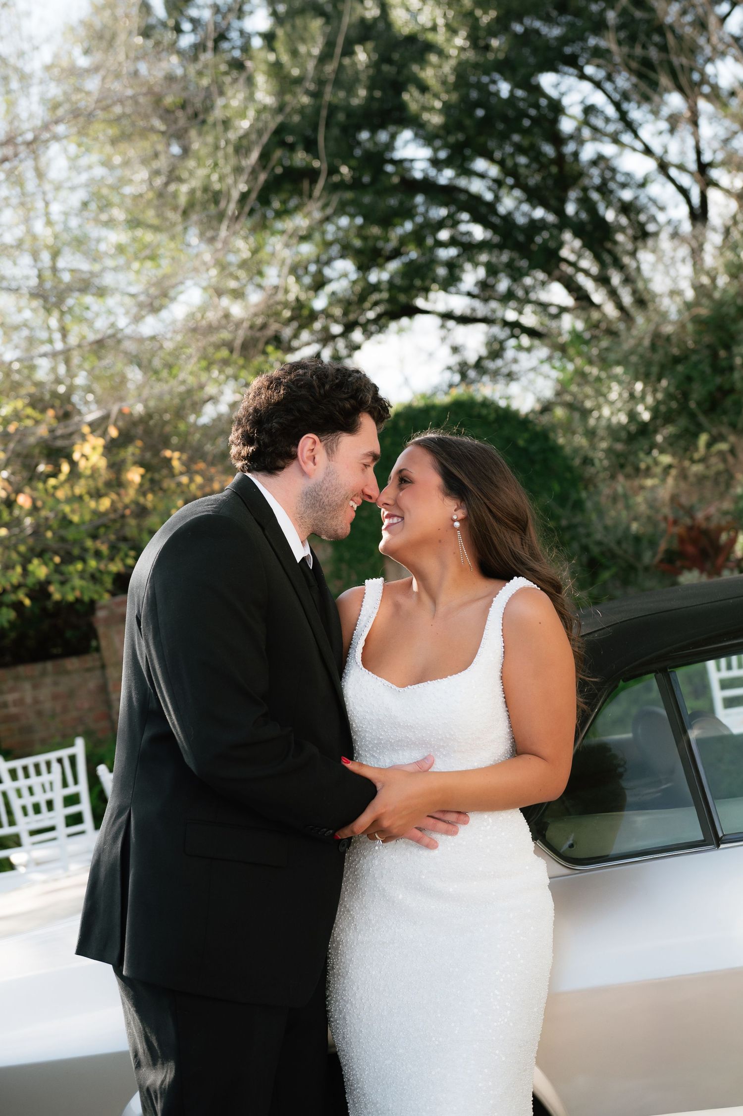 A bride and groom share a romantic moment, smiling at each other in formal wedding attire outdoors.