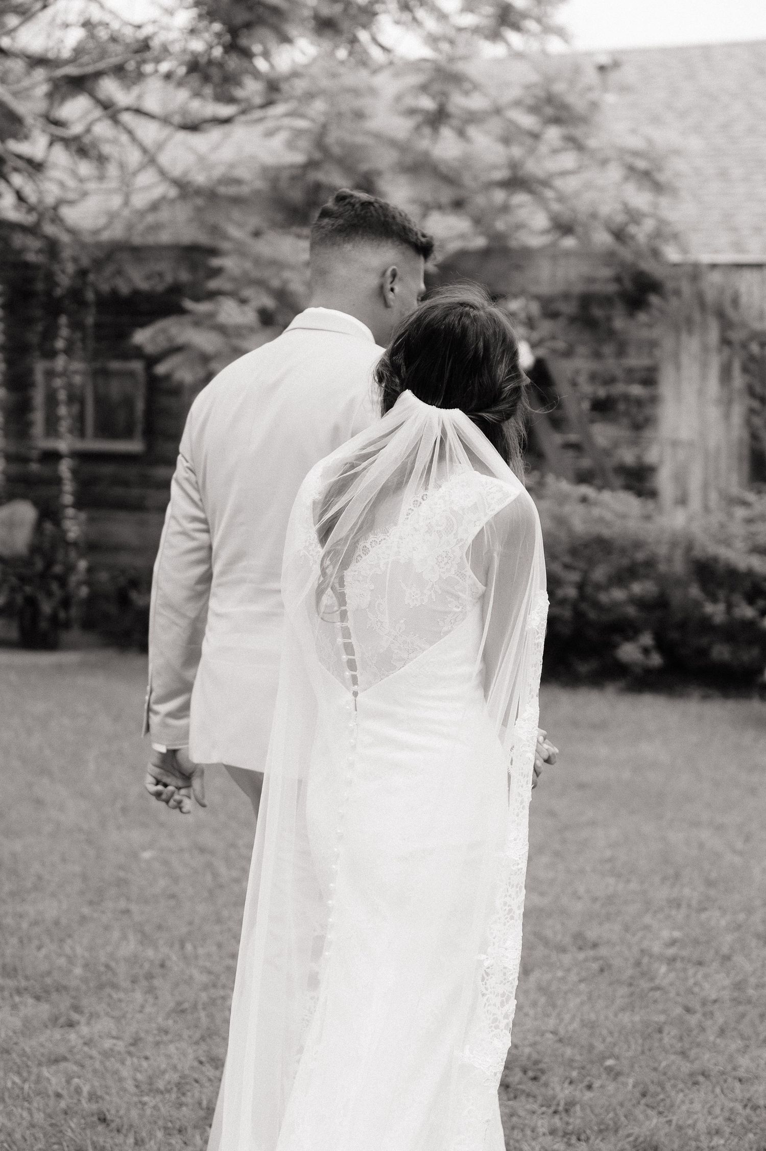 Black and white photo of bride and groom holding hands, viewed from behind, standing on green lawn near rustic building.