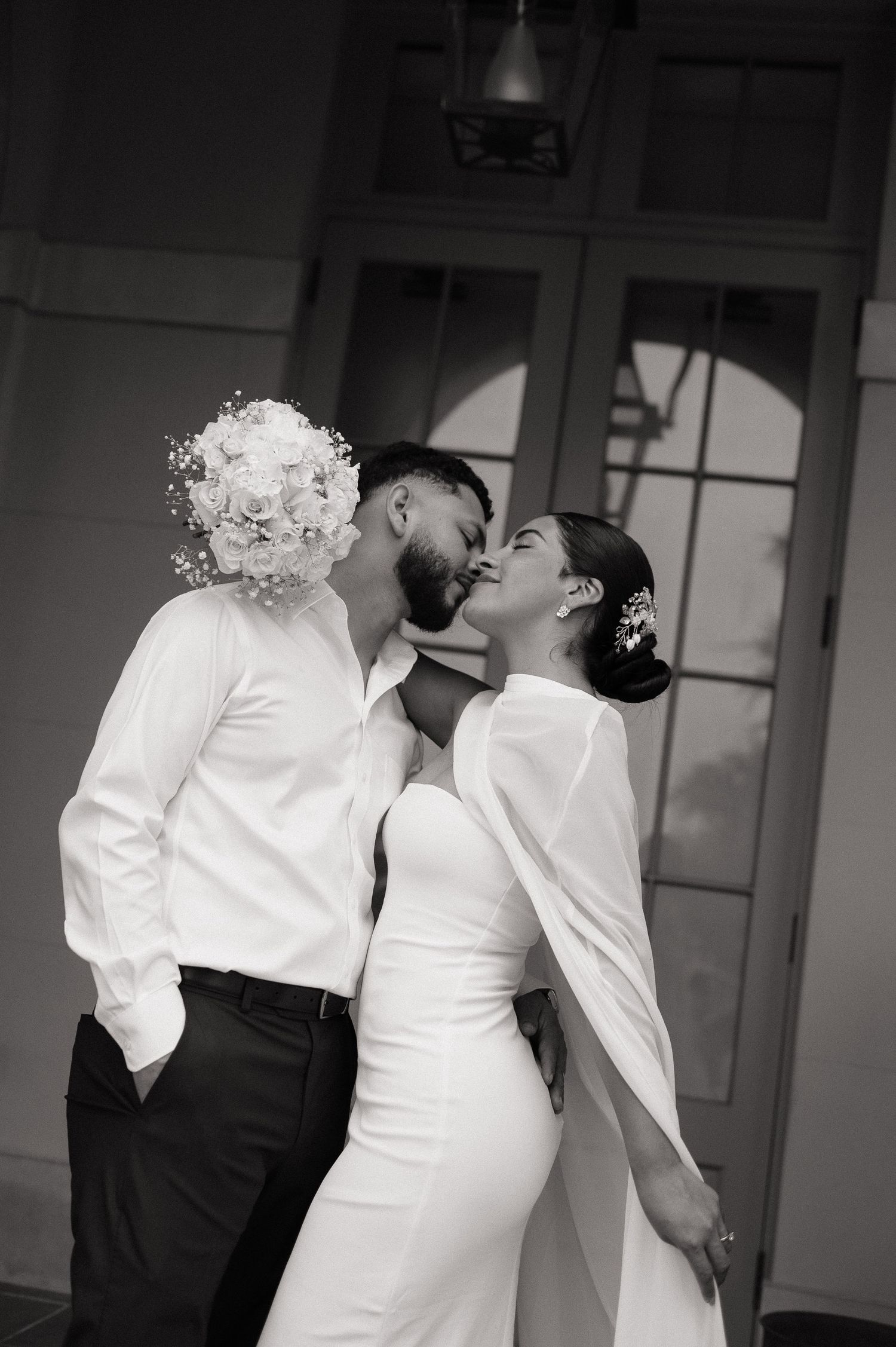 Black and white photo of a bride and groom sharing a romantic kiss, bride holding a white bouquet in an elegant wedding gown.