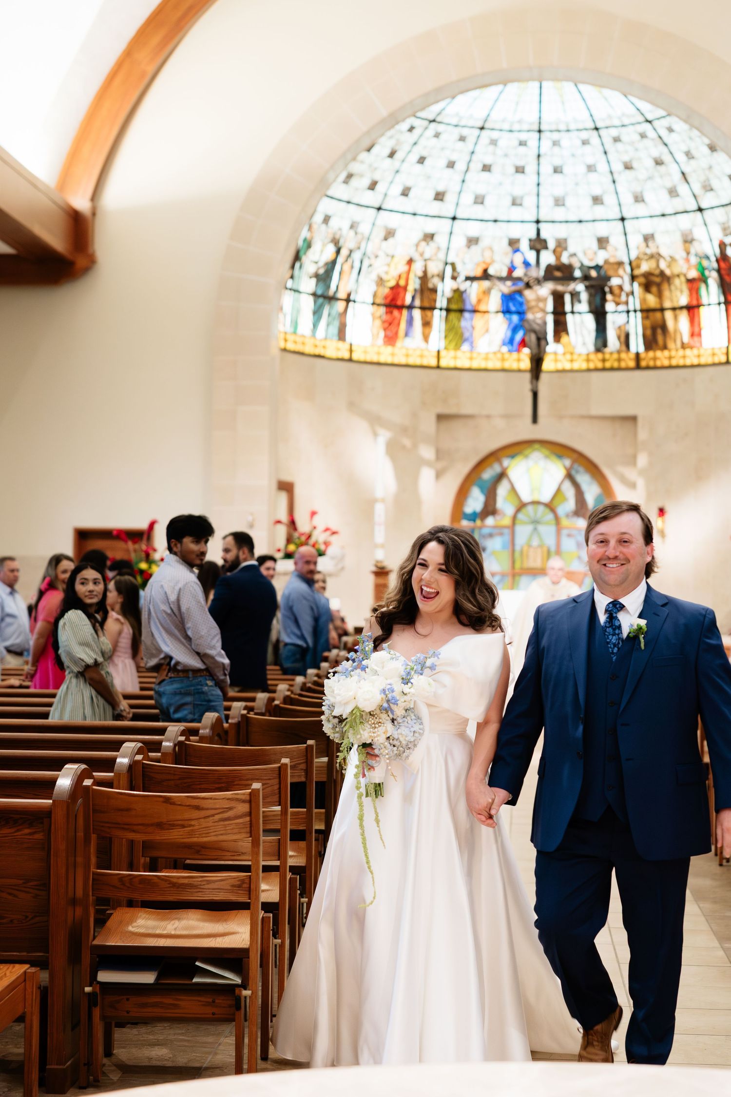 Bride and groom walking down church aisle smiling with stained glass dome backdrop.