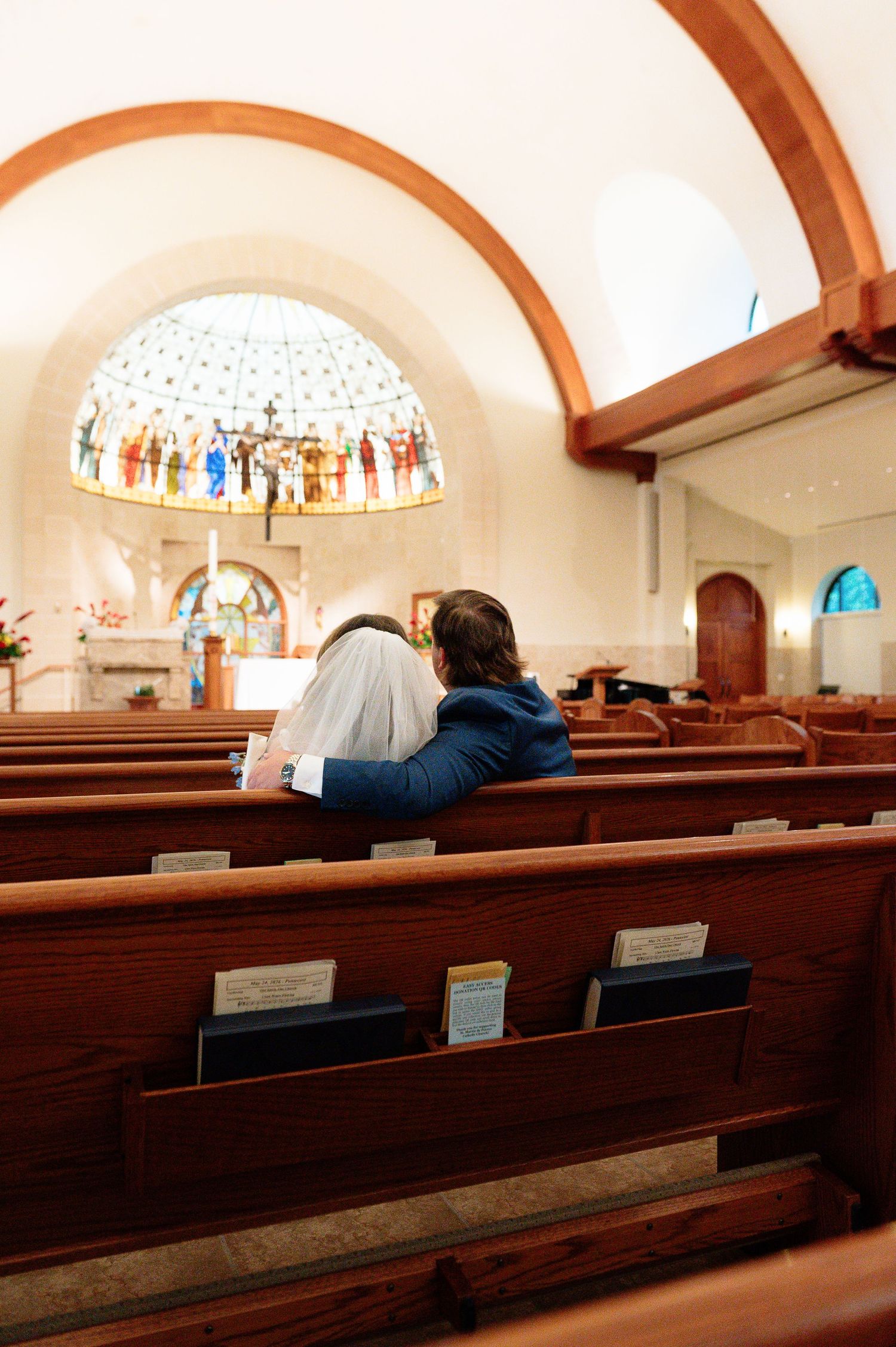 Bride and groom sitting together in church pews before a beautiful stained glass window altar.