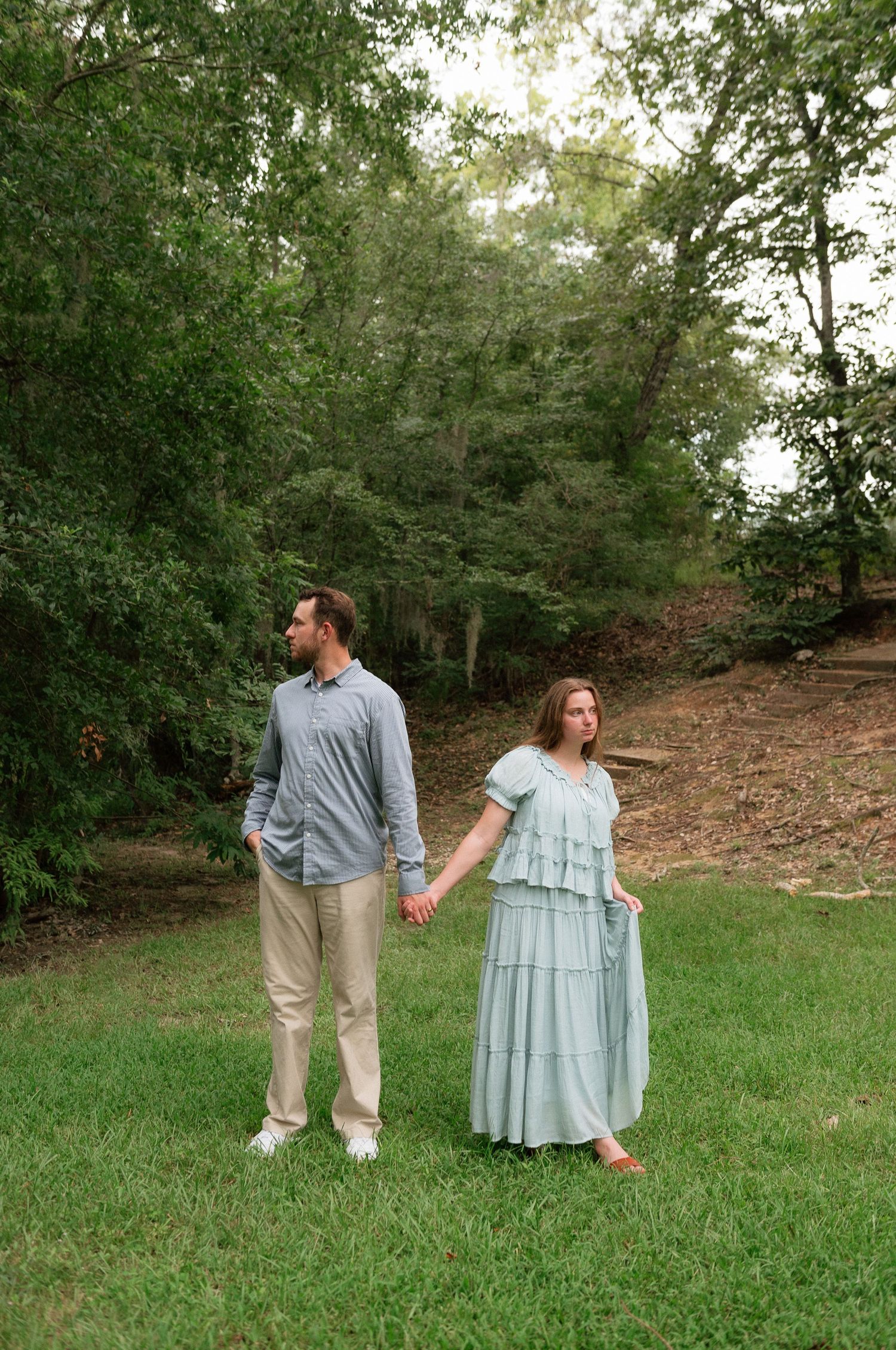A couple holds hands in a lush green yard, man in blue shirt and khakis, woman in a flowing mint dress.