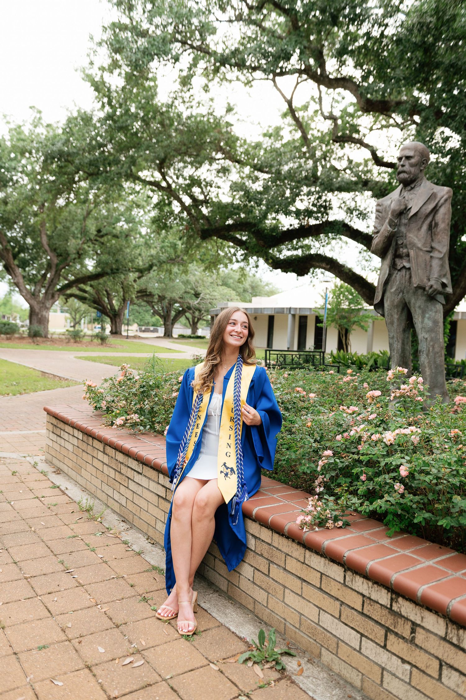A smiling graduate in blue regalia sits on a brick wall surrounded by roses near a bronze statue on a university campus.