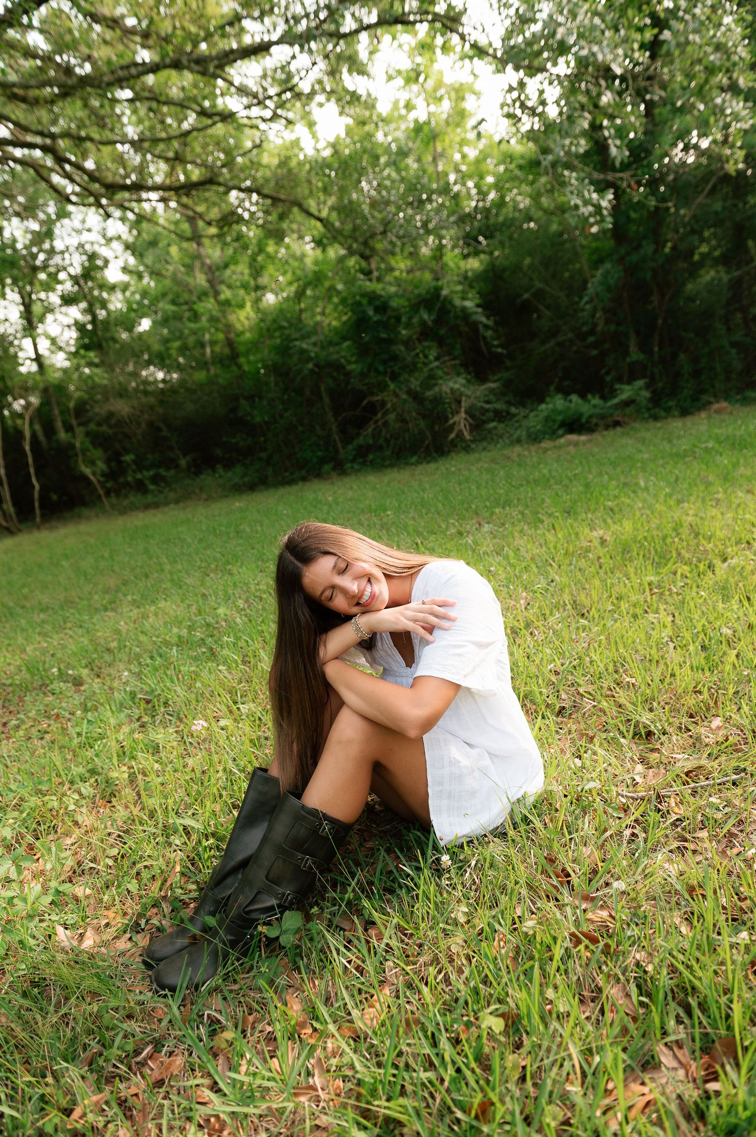 Young woman in white dress and black boots sitting on green grass, smiling, with lush trees in the background.