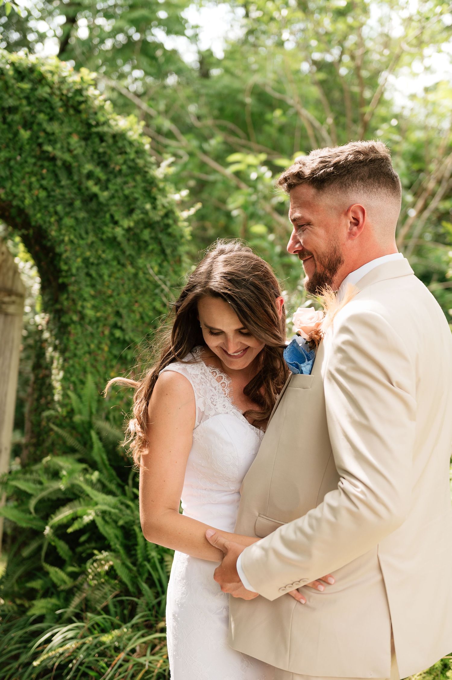 Smiling bride and bearded groom in cream suit embrace in lush garden with ivy-covered arch backdrop.