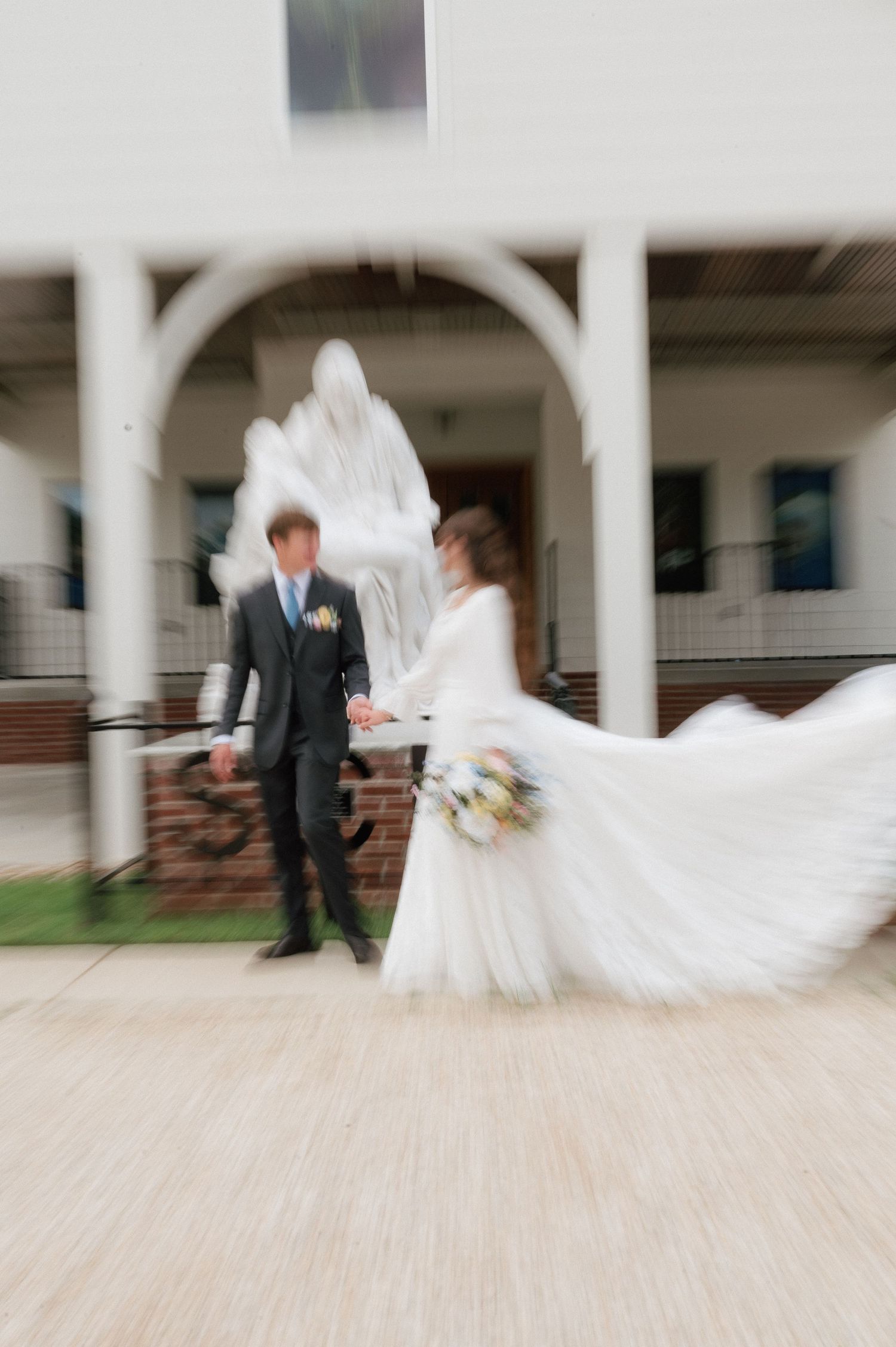 Bride and groom hold hands outside a church with a white statue, bride's gown flowing in motion blur.