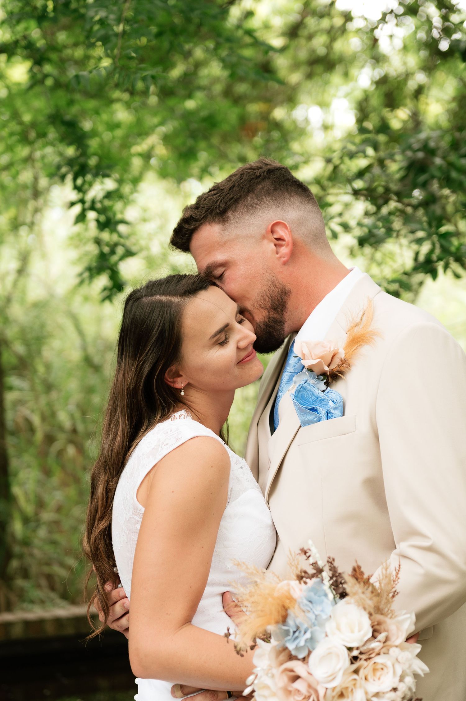 Bride and groom share a tender forehead kiss outdoors, she holds a boho bouquet, he wears a tan suit with blue bow tie.