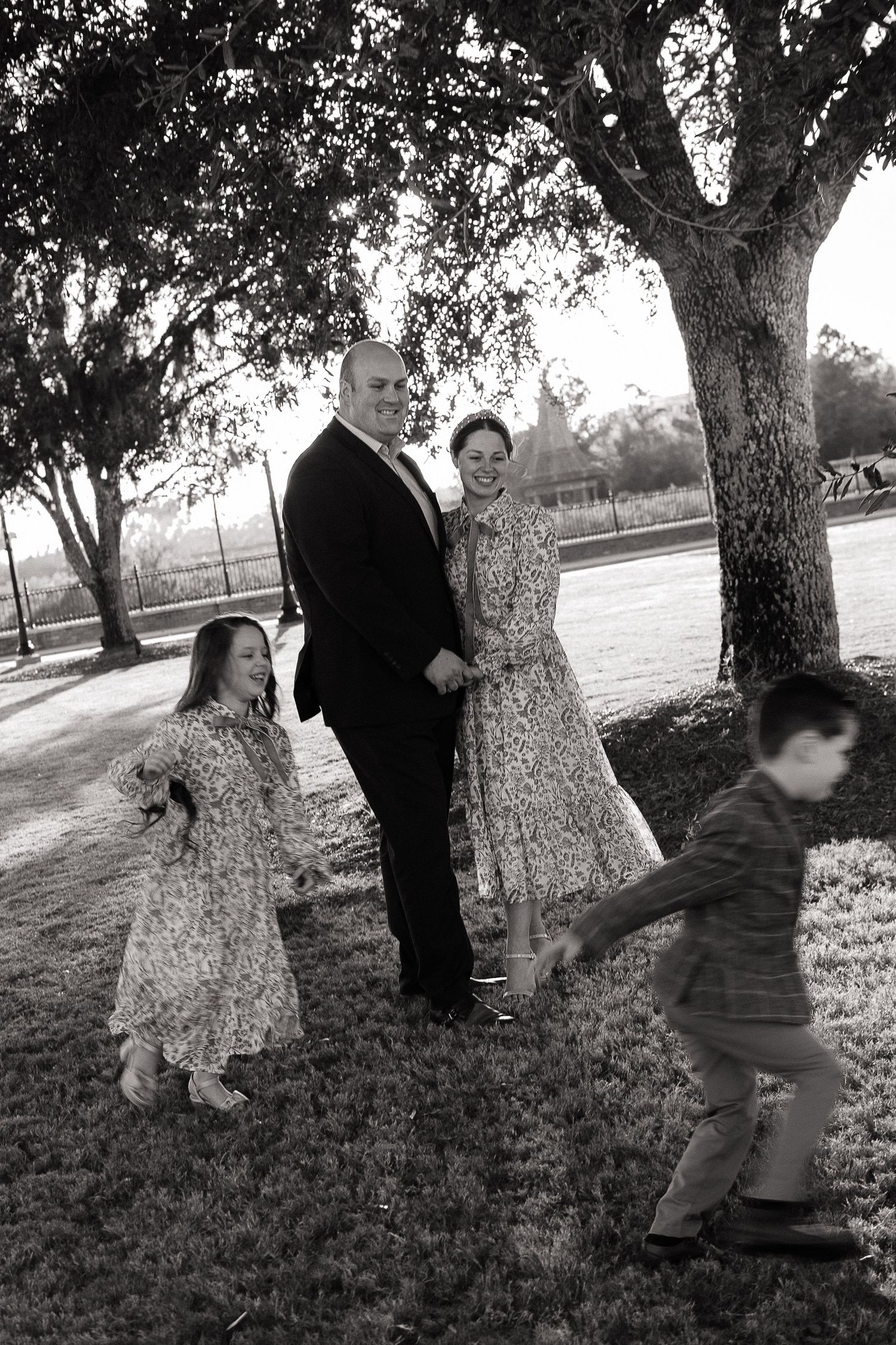 Black and white family photo of parents with two young children playing under trees in a park setting.