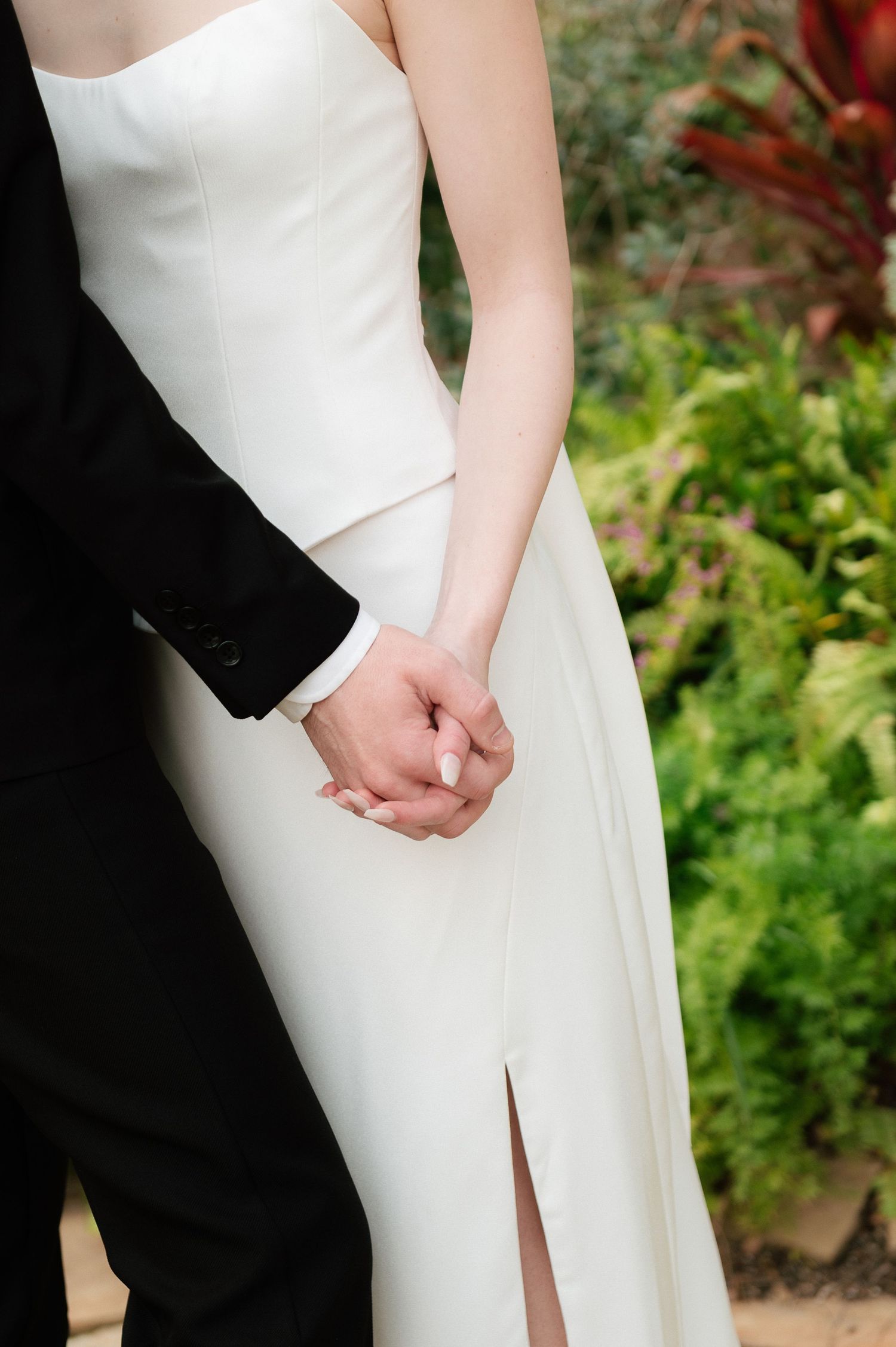Bride and groom holding hands, wearing white wedding gown and black tuxedo, with lush green garden backdrop.