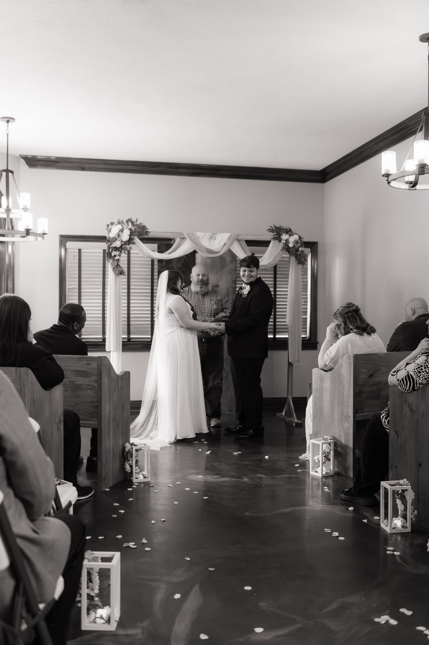 Black and white wedding ceremony photo of bride and groom holding hands at the altar with guests seated in pews.
