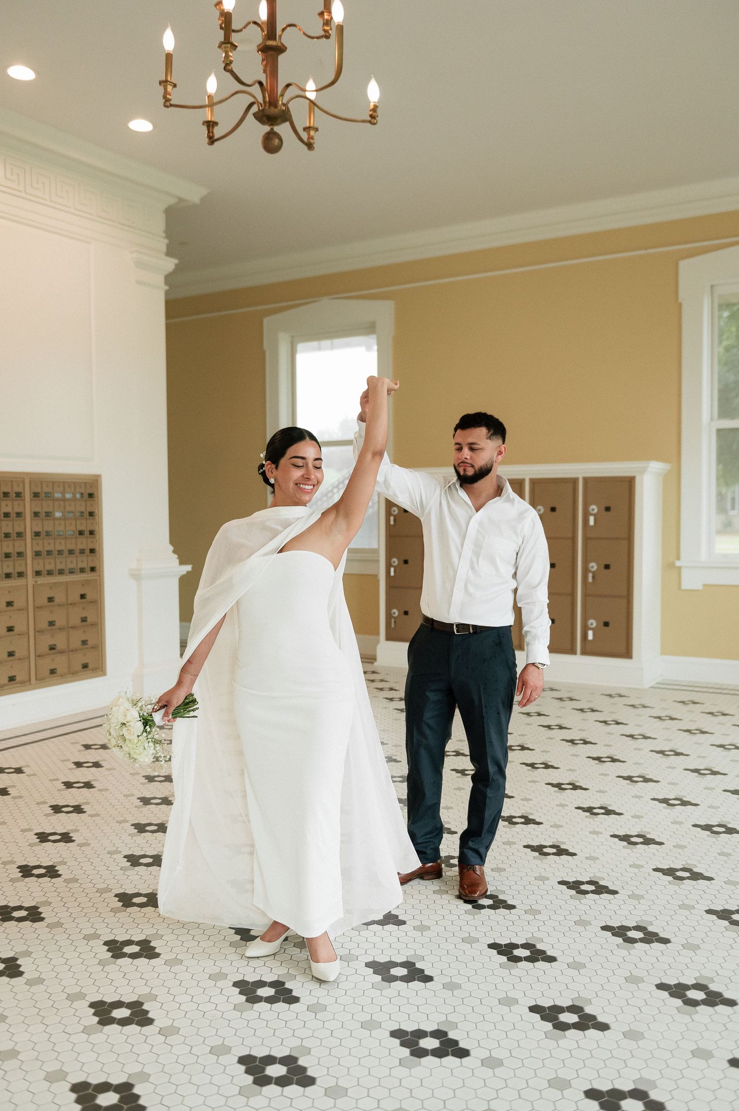 Bride and groom dancing in elegant white tiled lobby with chandelier on their wedding day.