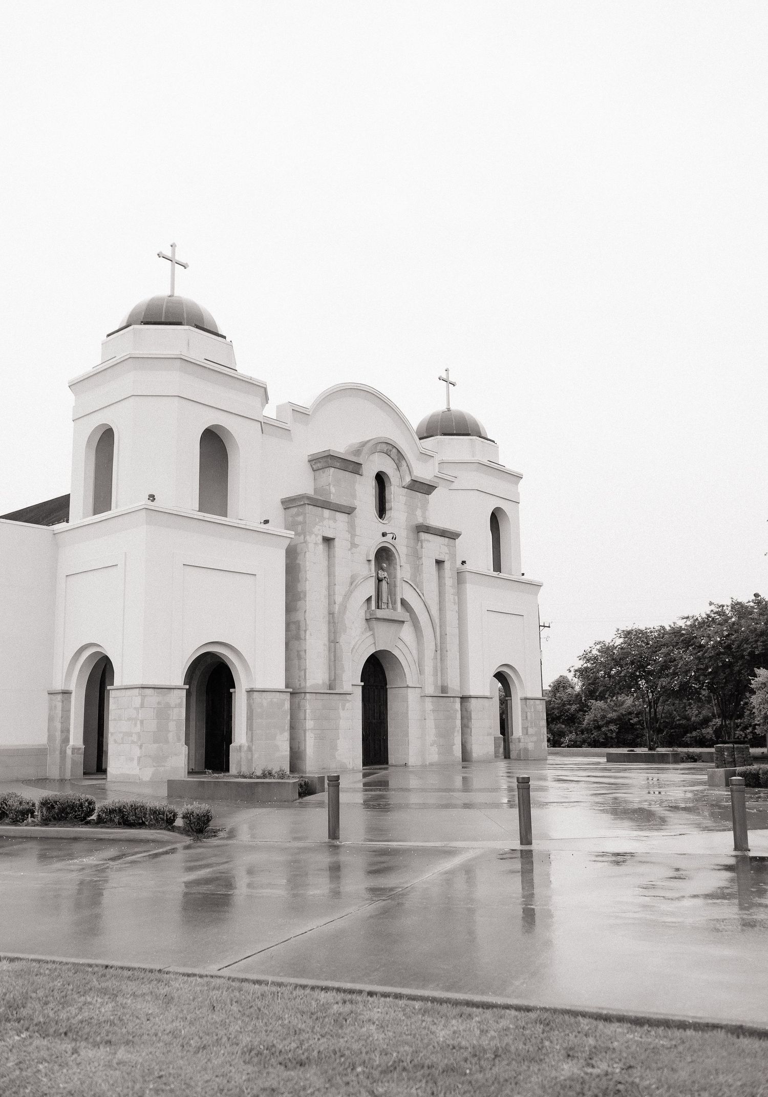 Black and white photo of white Spanish-style church with domed towers and cross on overcast rainy day.