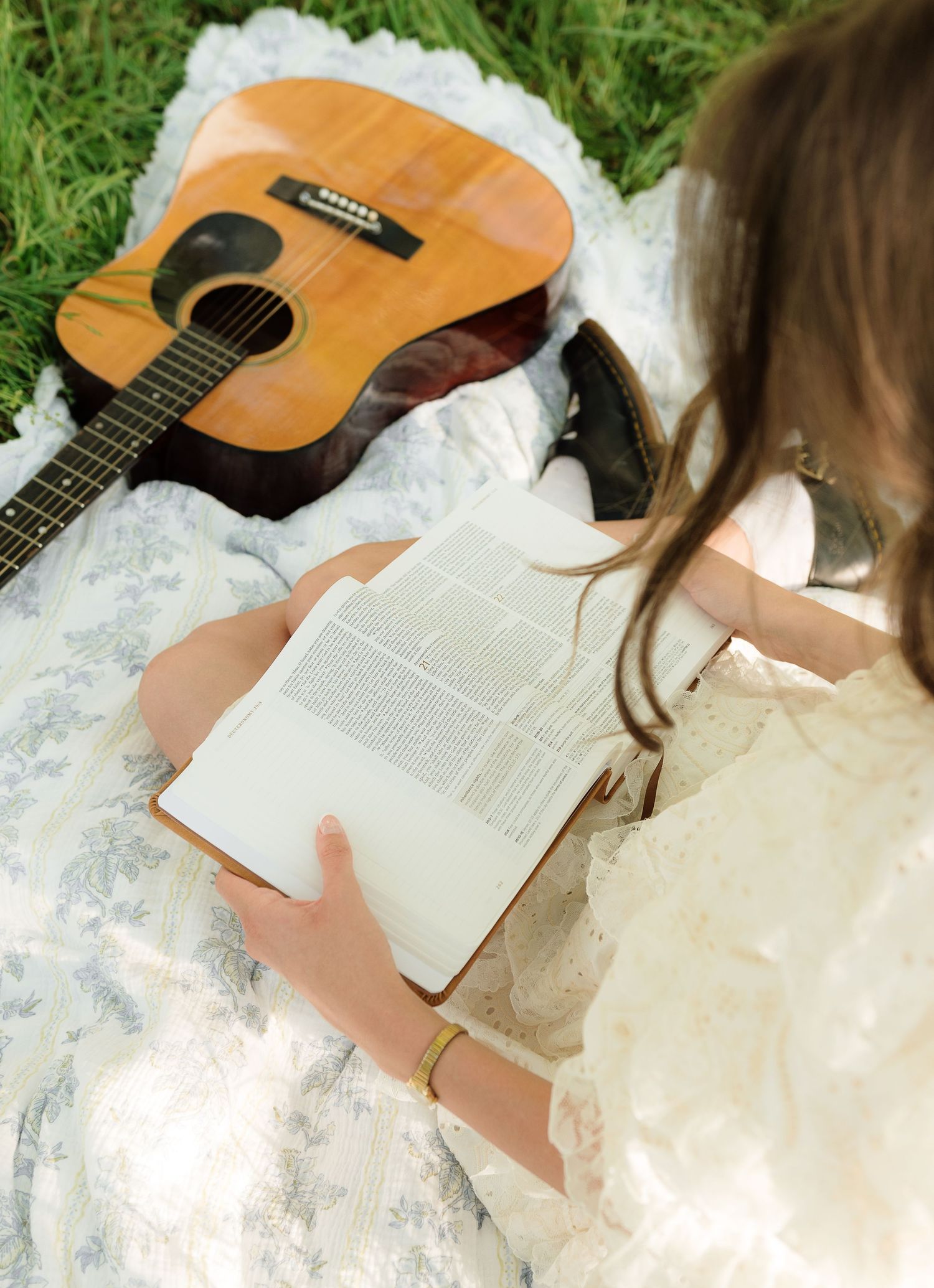 Young woman in white dress reading a book on a blanket in grass beside an acoustic guitar.