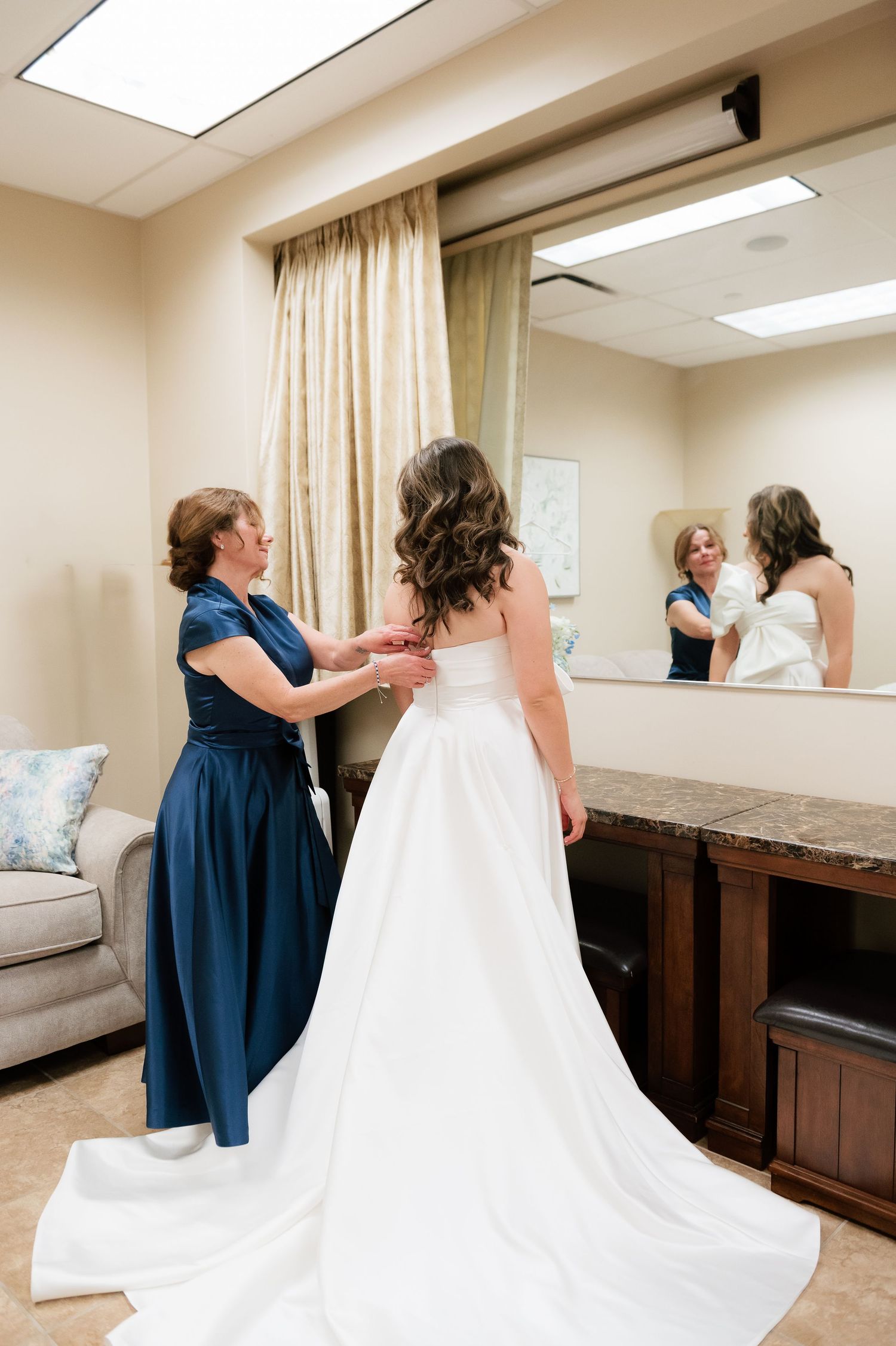 A bride in a white gown gets her dress fastened by a woman in navy blue, reflected in a large mirror.